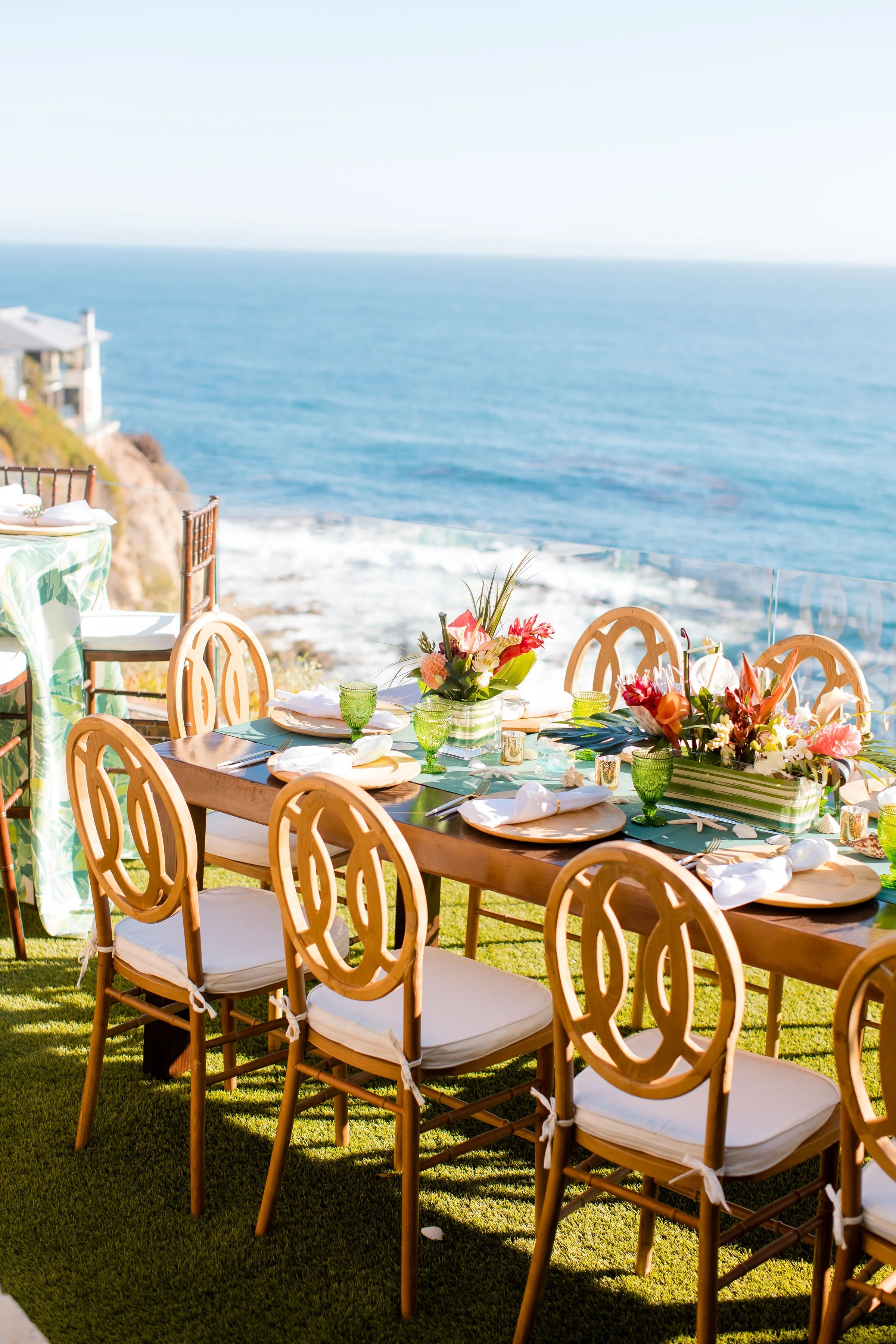 Elegant oceanfront event tables cape with fresh floral arrangements, green glassware, and coastal views.