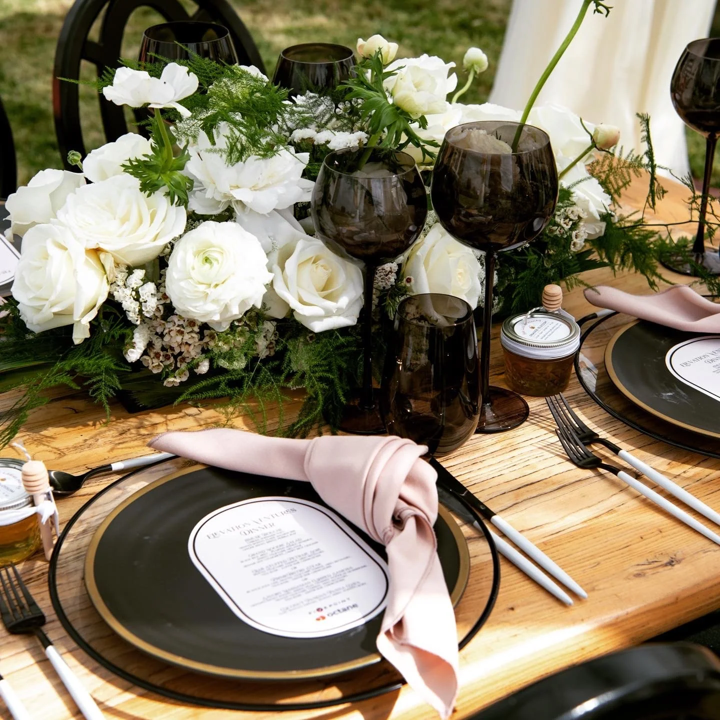 A table decorated with white flowers and greenery, set for a meal with black and gold plates, pink napkins, and black glasses. A menu is placed on each plate, and cutlery is arranged on the sides of the plates.