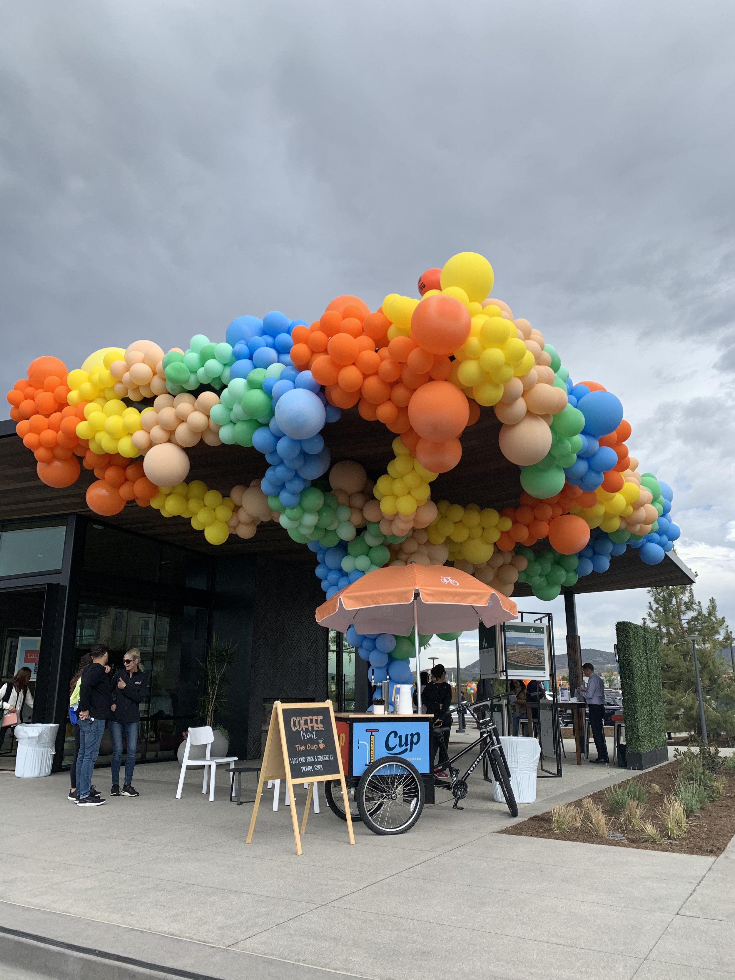 Colorful balloons decorating the roof of a modern building, with a group of people gathered outside. There is a coffee stand with a chalkboard sign, a bicycle, and an umbrella in front of the building, overcast sky in the background.