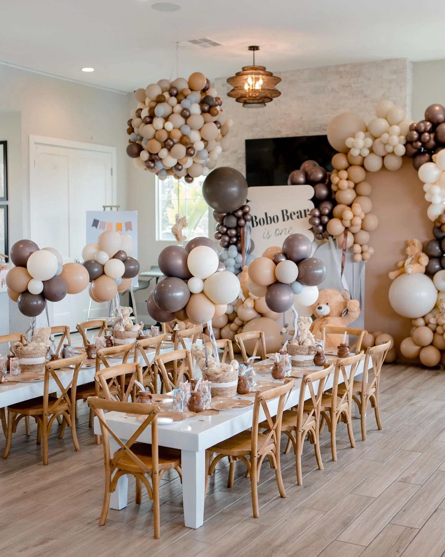 Elegant indoor first birthday celebration featuring neutral balloon garlands, wooden dining chairs, coordinated table settings, and custom party decorations.