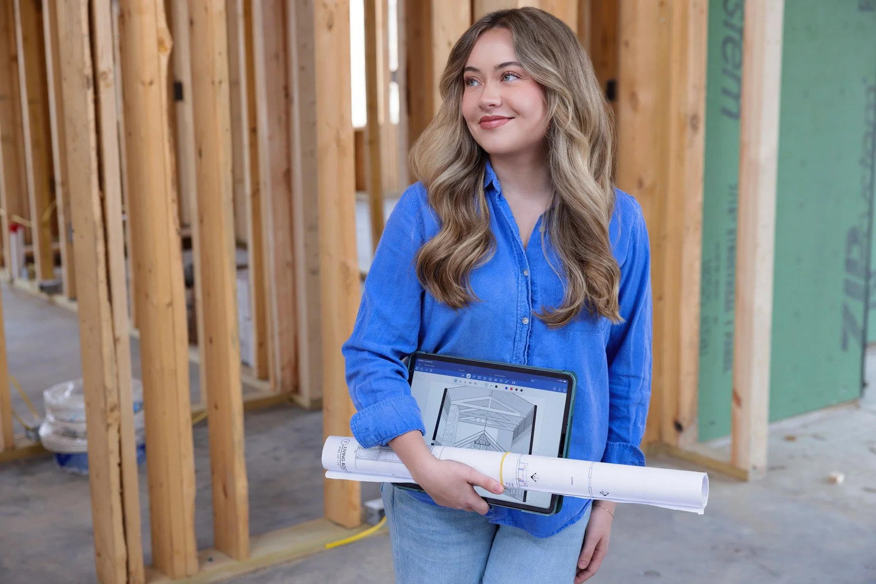 Young woman in a construction site holding blueprints and a tablet displaying a 3D building design.