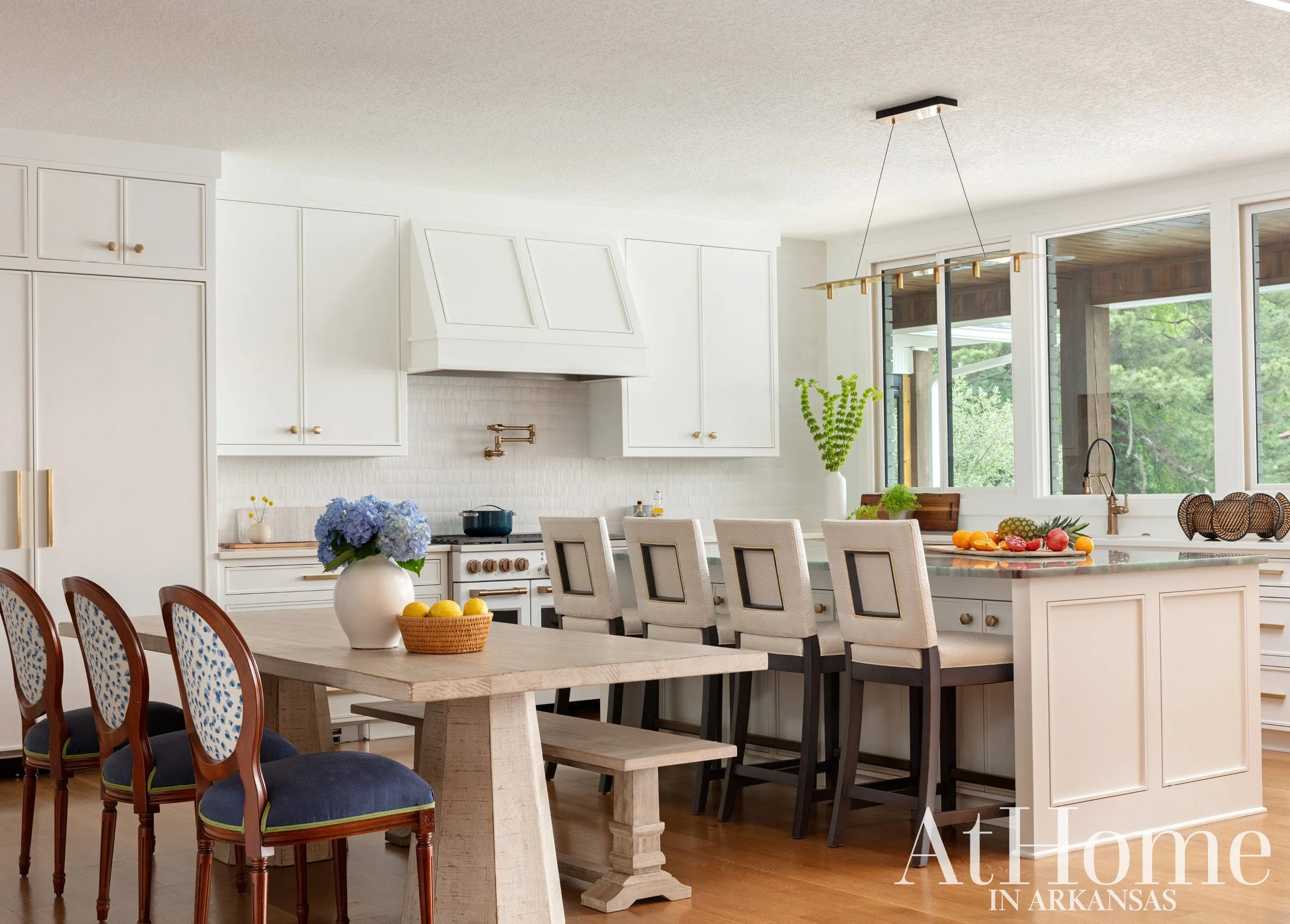 Kitchen with white cabinets, a central island with bar stools, a dining table with eight chairs, and large windows showing greenery outside.