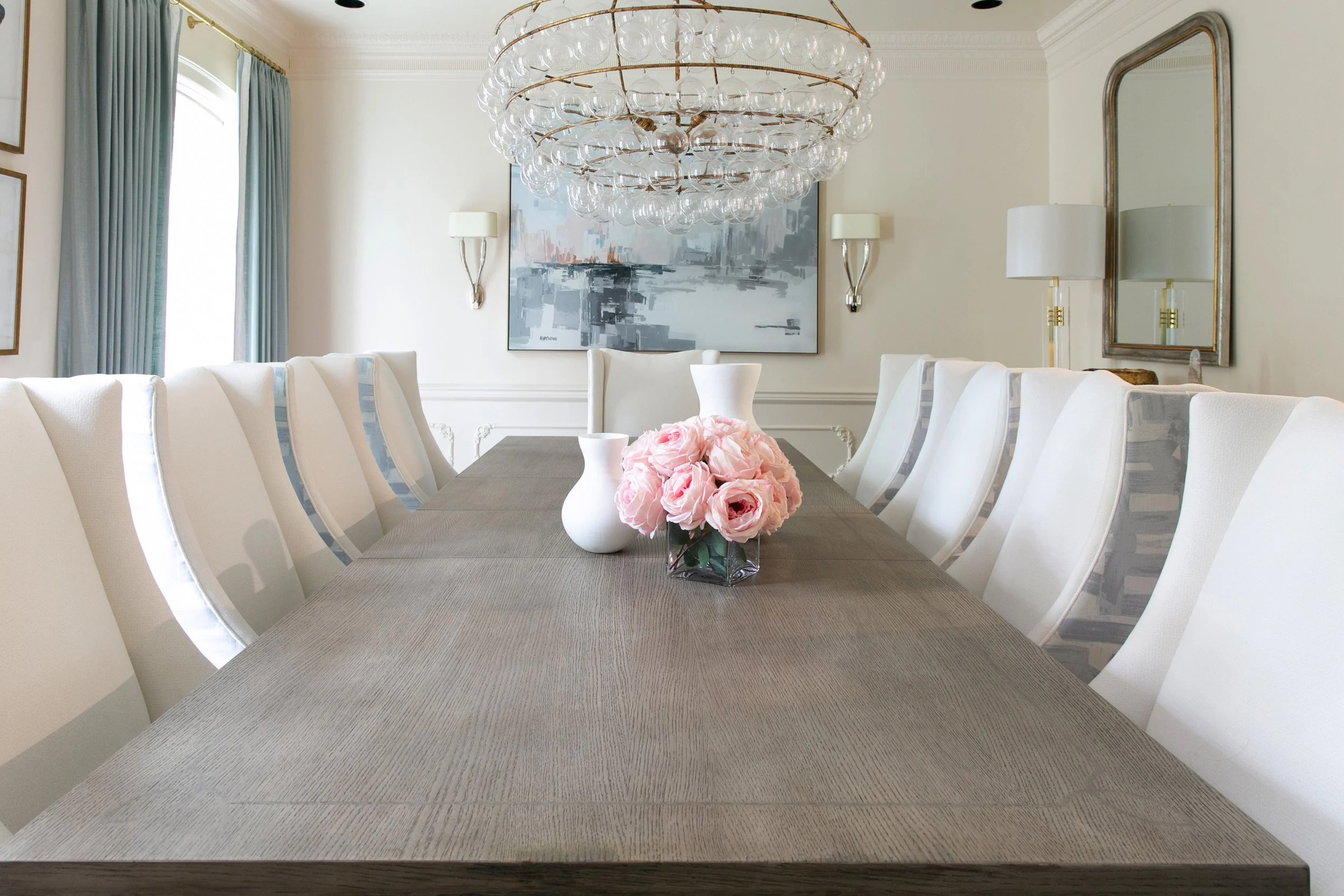 A dining room with a long wooden table, surrounded by white upholstered chairs, a chandelier hanging above, pink flowers and white vases on the table, and artwork on the wall.