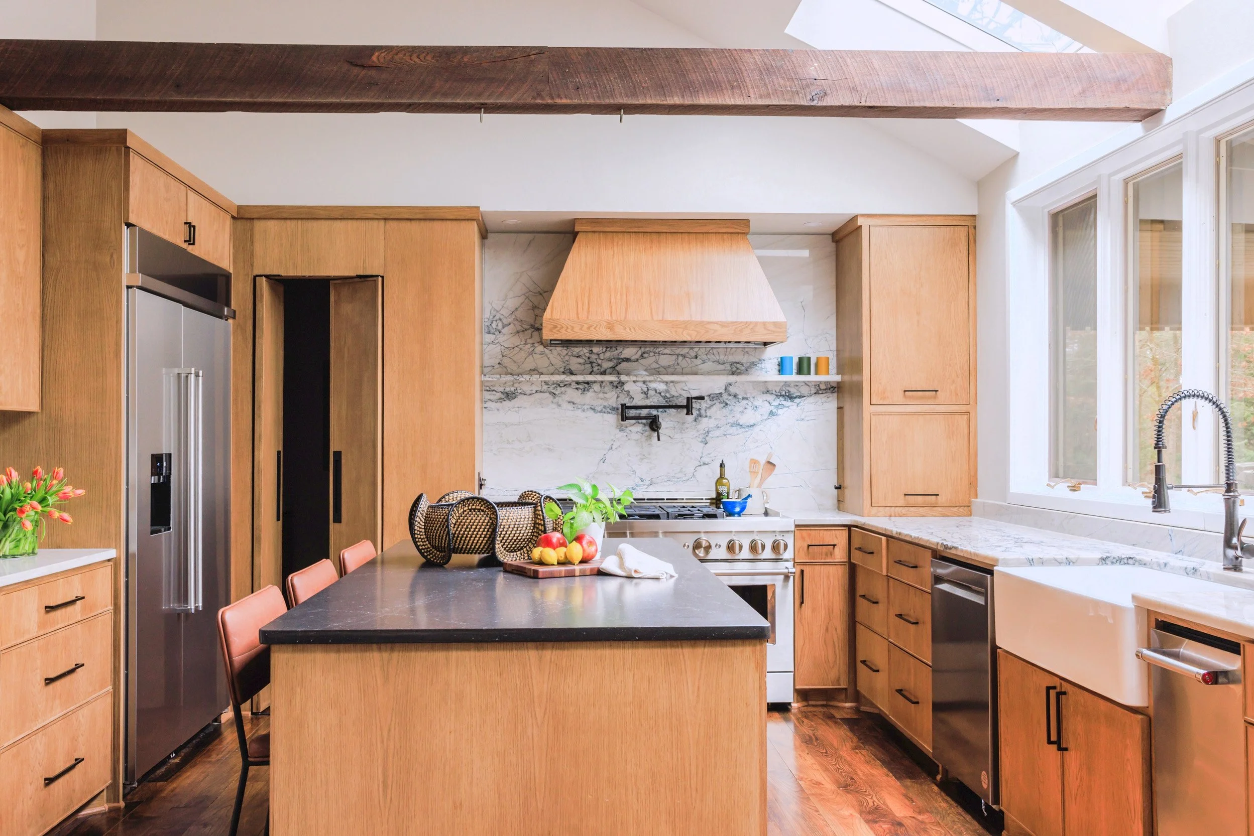 Kitchen with wooden cabinets, a black countertop island with chairs, a stainless steel refrigerator, a marble backsplash, and a farmhouse sink near large windows.