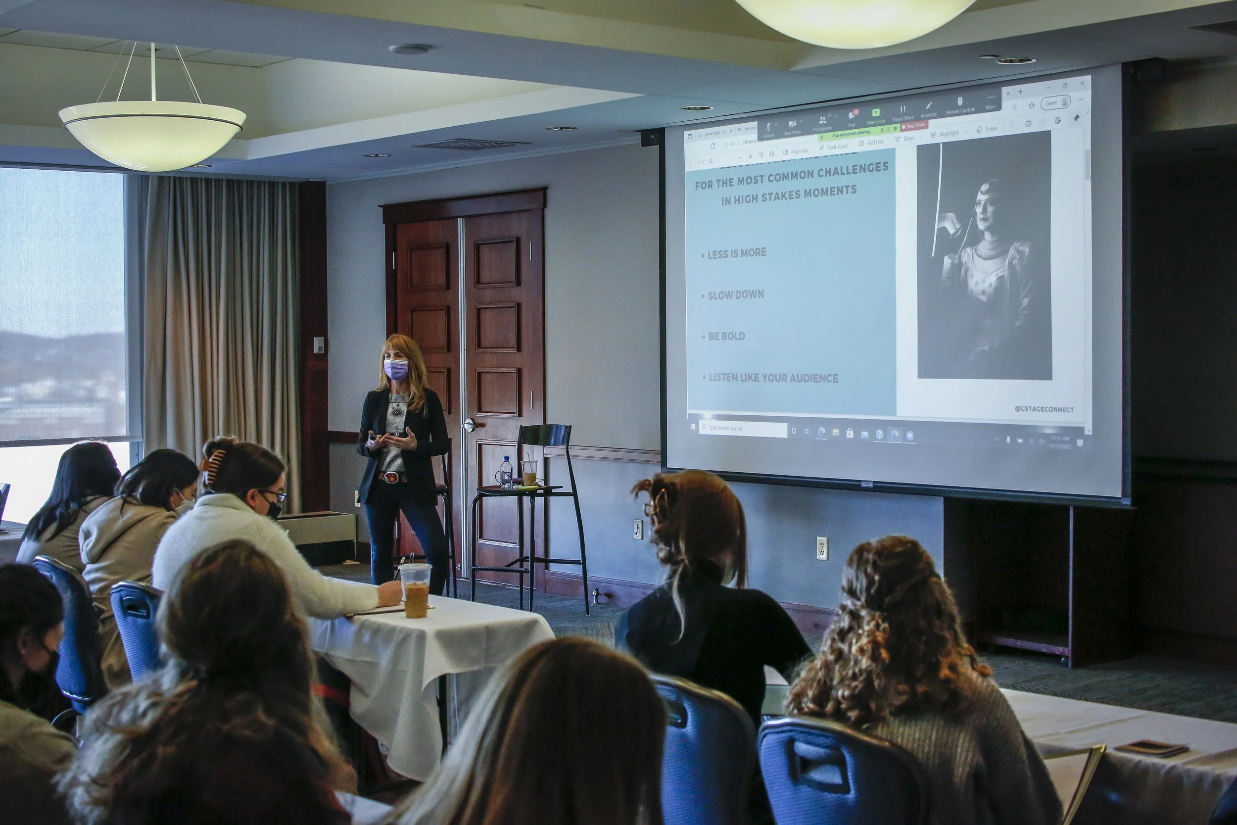 A woman giving a presentation on stage at a conference room with a large projector screen displaying a PowerPoint slide. The slide contains text about challenges in high-stakes moments and an image of a woman in traditional attire. Audience members are seated at tables, some taking notes.