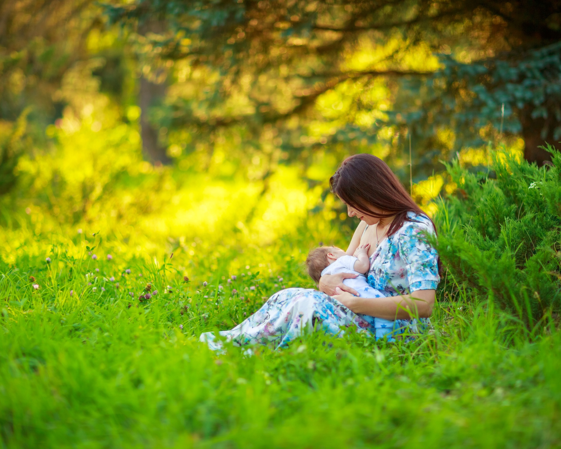 Image of woman in green grass with forest behind her in a floral dress nursing a baby.