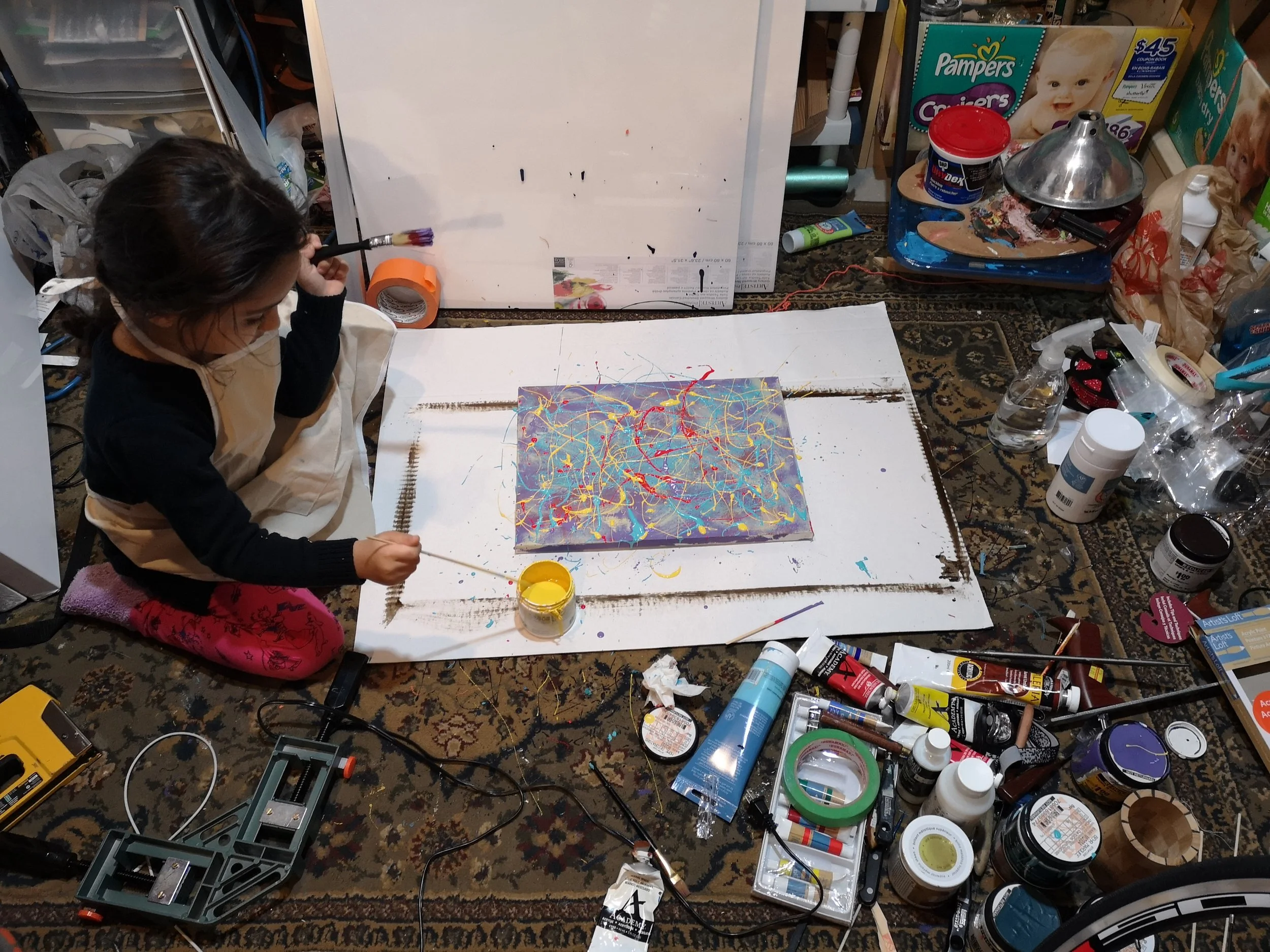 A young girl sits on a patterned rug, painting on a canvas with colorful splashes. The workspace around her is cluttered with paint supplies, brushes, and art tools, and there are boxes of diapers in the background.