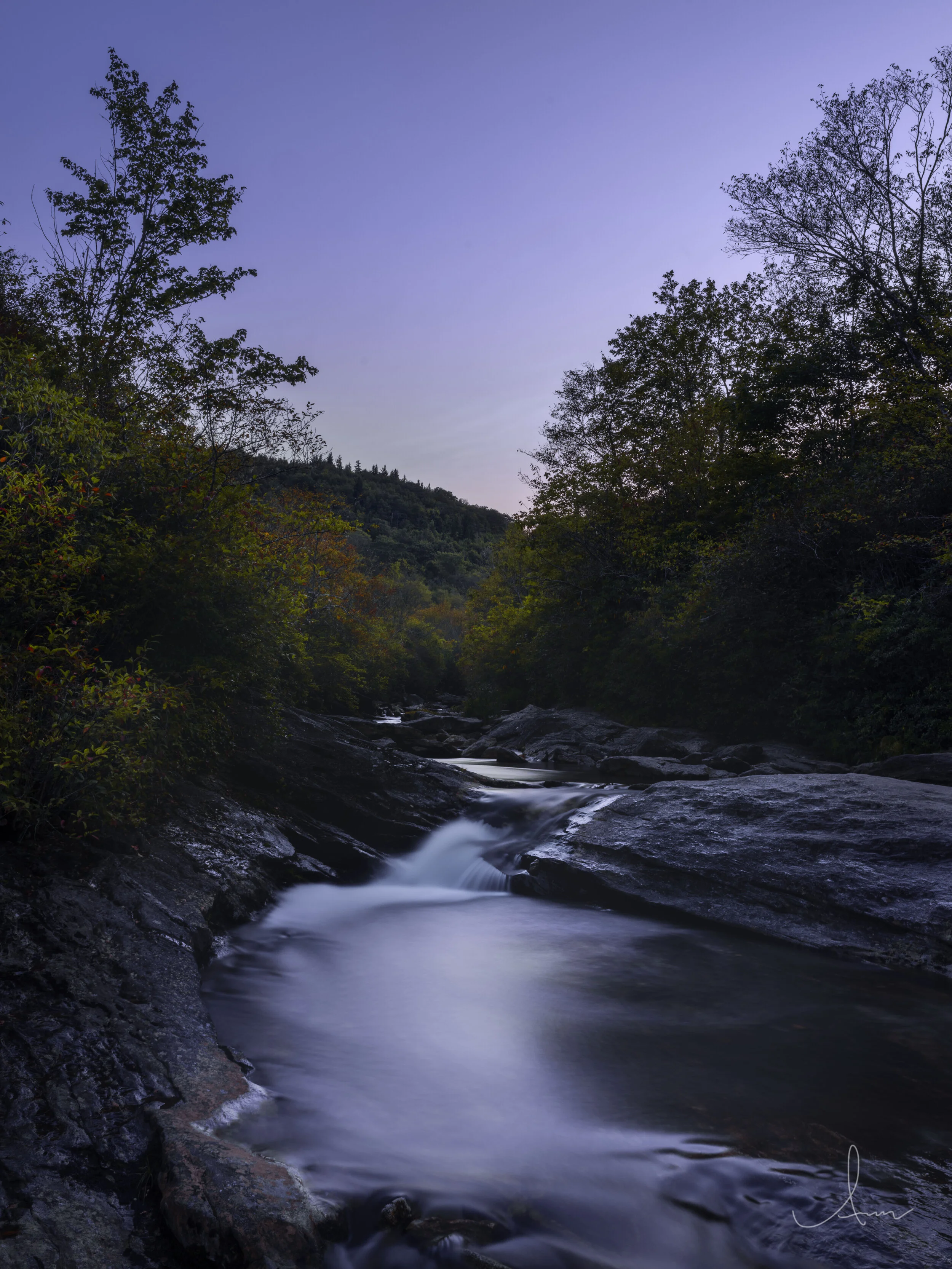 A tranquil river flowing through a rocky landscape with surrounding trees under a twilight sky.