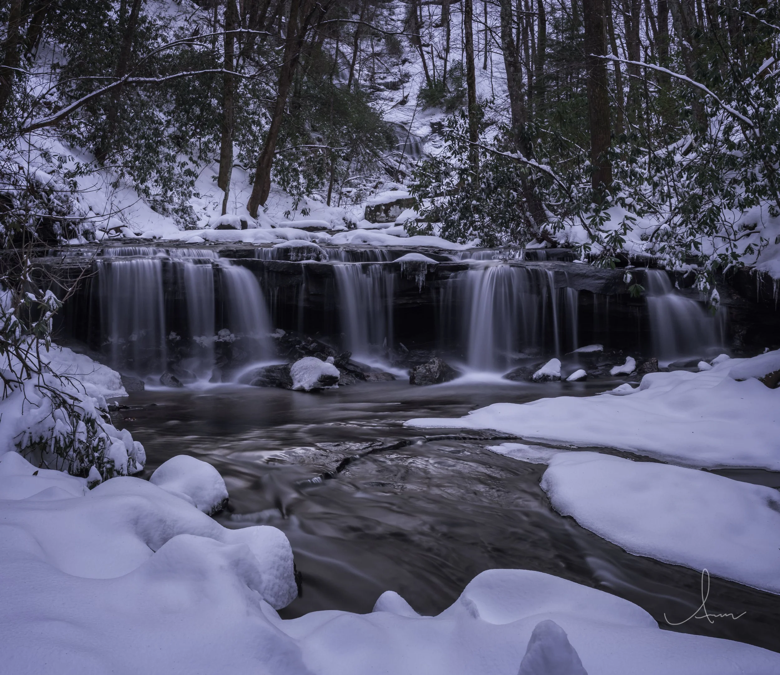 Snow-covered forest with a small waterfall cascading over rocks, surrounded by trees.