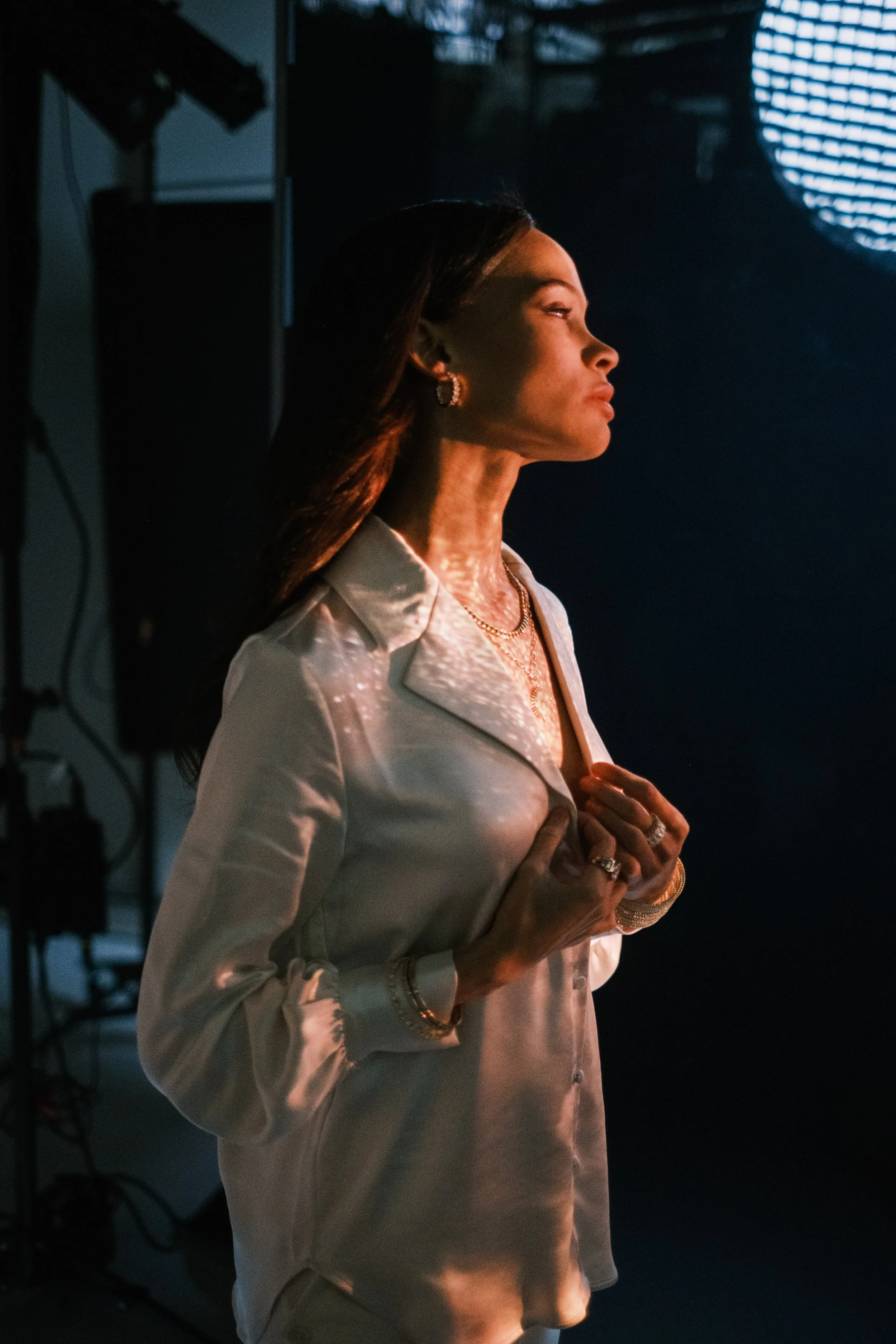 A woman with long dark hair, wearing jewelry, is standing in profile in front of a dark background with a large, textured light source to her side, creating shadows on her face.