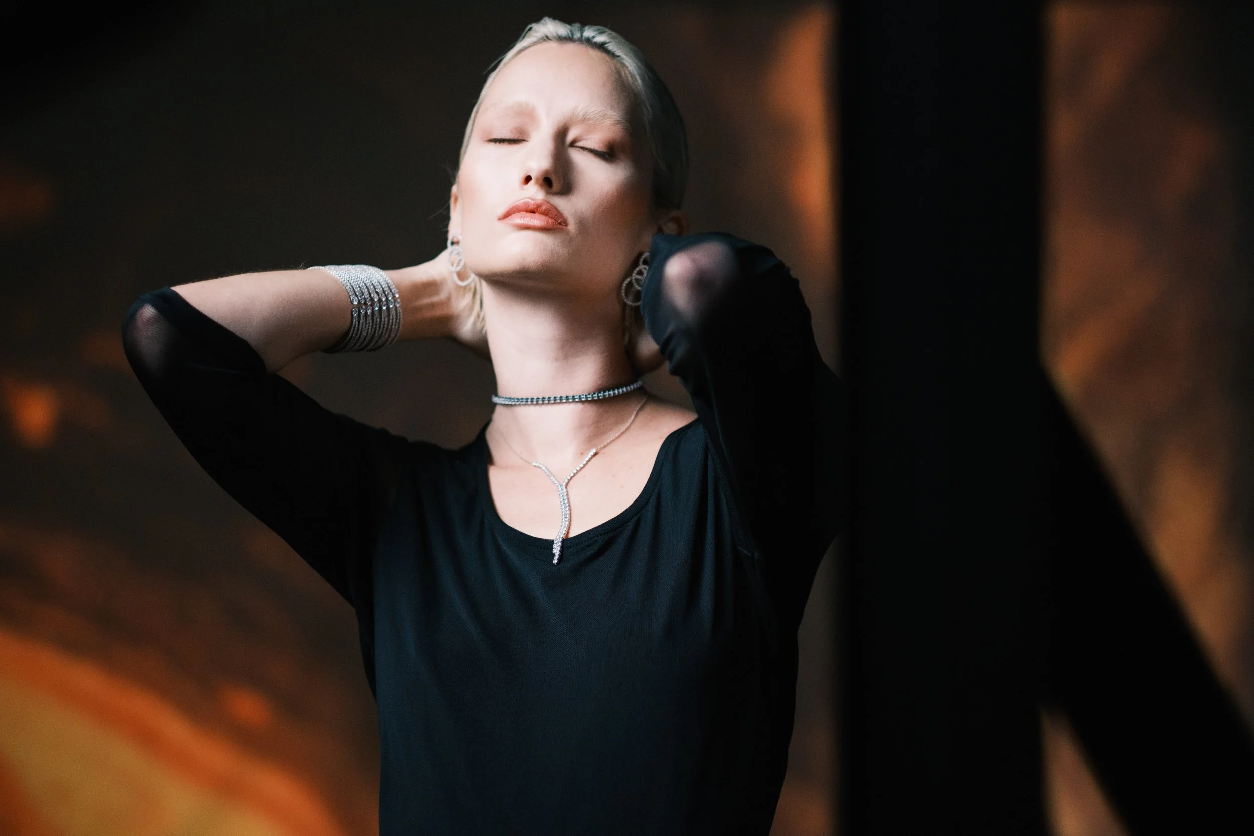A woman with platinum blonde hair and fair skin is pictured with her eyes closed, wearing a black top with sheer sleeves and silver jewelry including a choker, earrings, bracelet, and necklace. She appears to be in a moment of relaxation or enjoyment