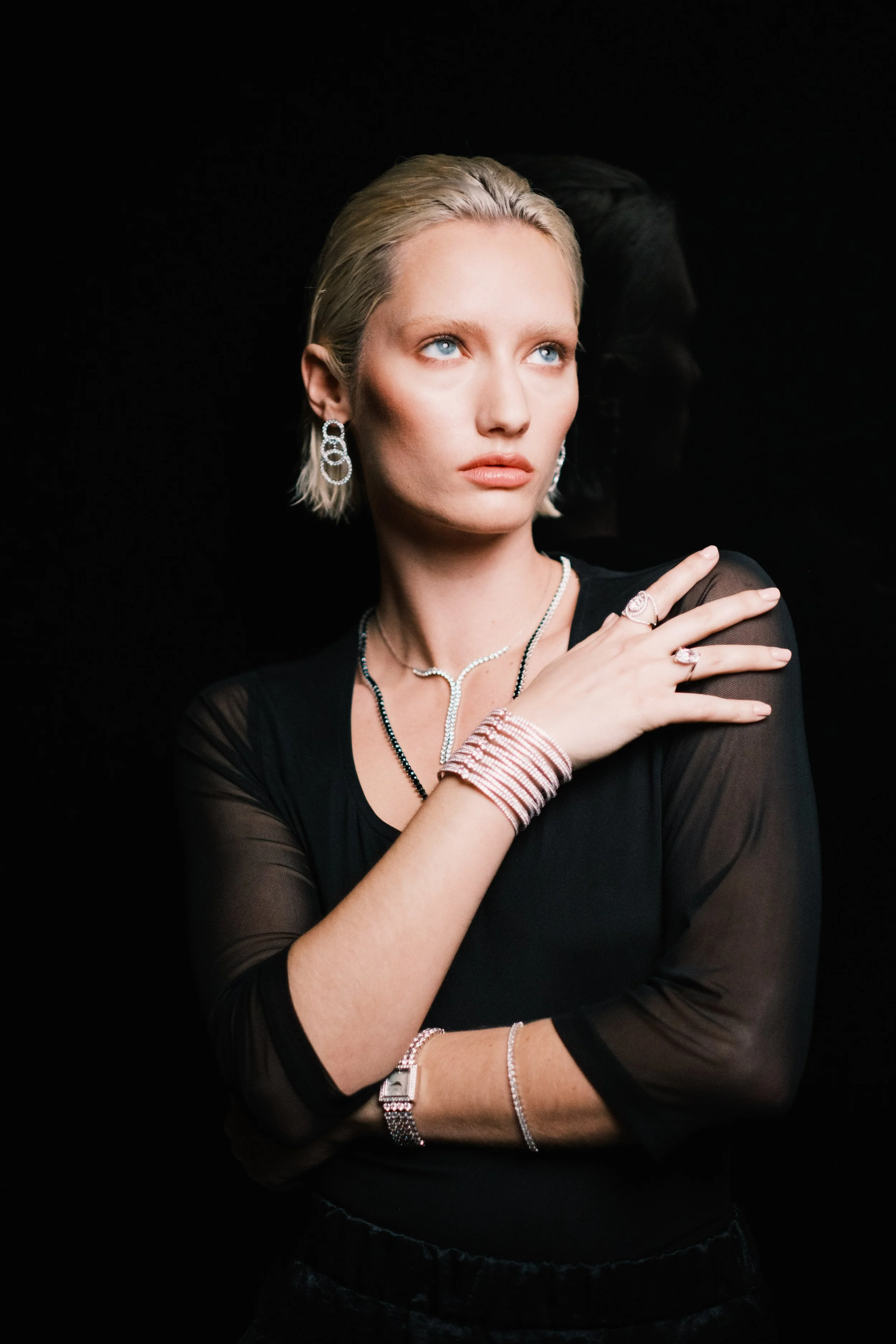 A woman with short blonde hair and blue eyes standing against a black background, dressed in black with silver jewelry, looking off to the side.