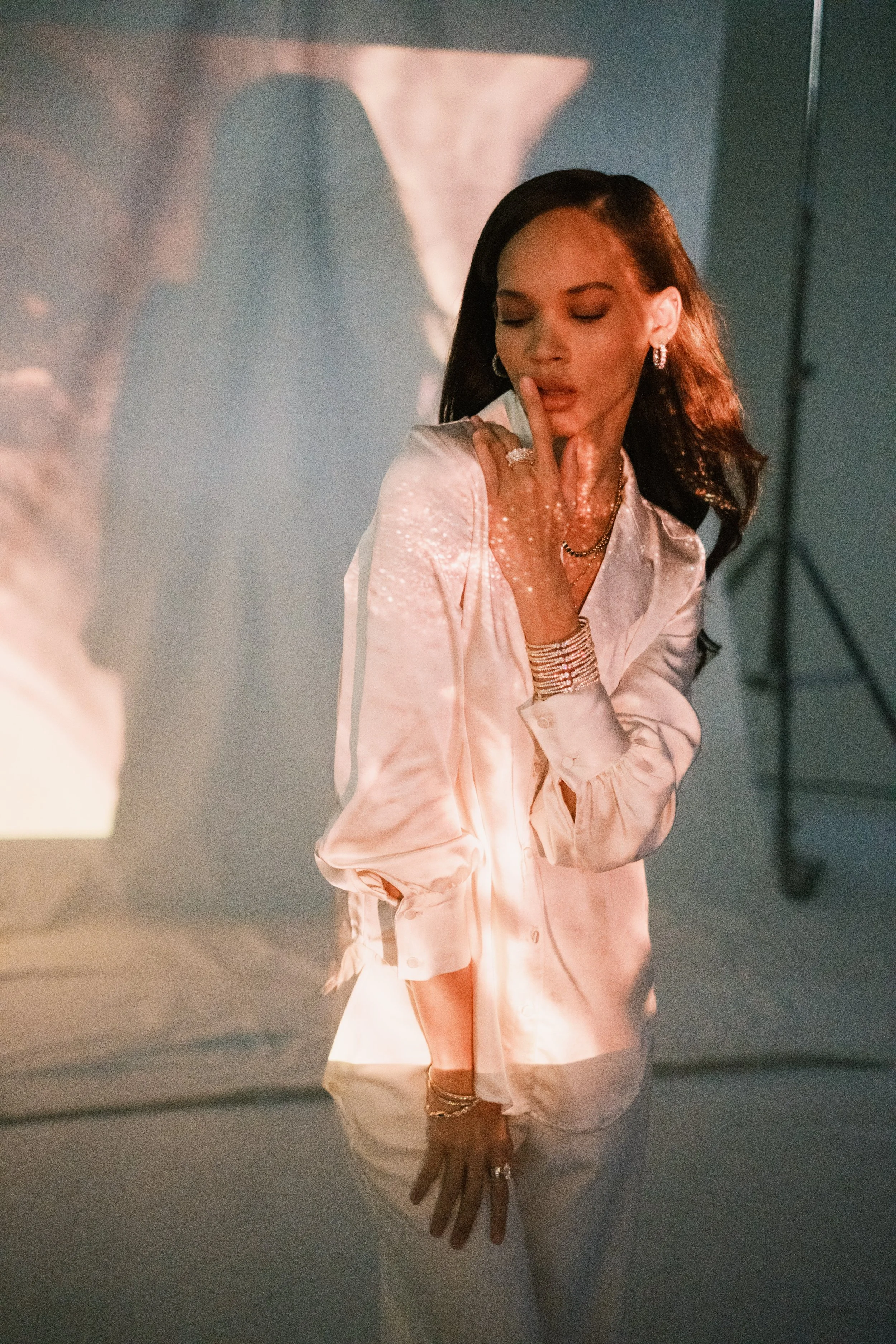 A woman with dark hair styled in waves, wearing a white satin blouse and multiple jewelry pieces, stands with her eyes closed and finger touching lips amid a soft, artistic lighting setting.