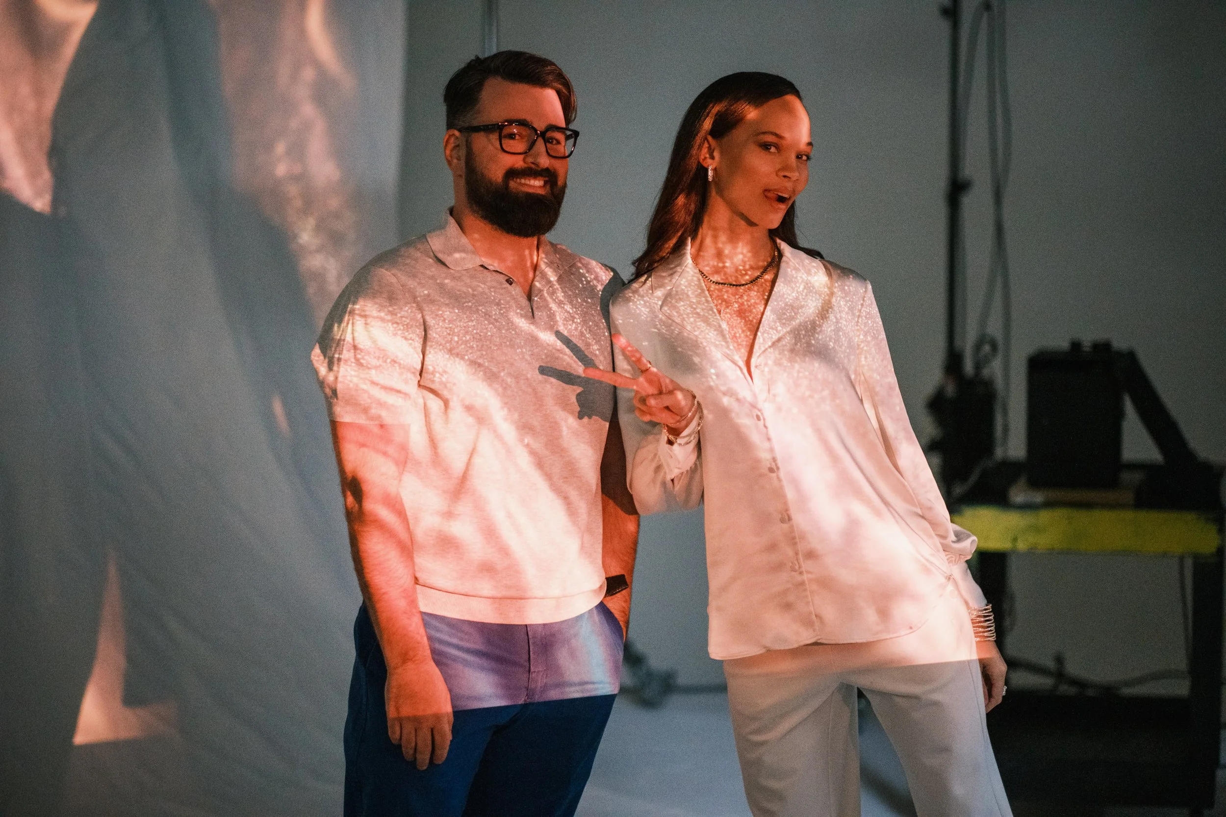 Two people, a man and a woman, posing together in front of a projected screen. The man has dark hair, a beard, glasses, and is wearing a light-colored polo shirt and dark pants. The woman has long, dark hair, and is wearing a shiny, satin-like blouse