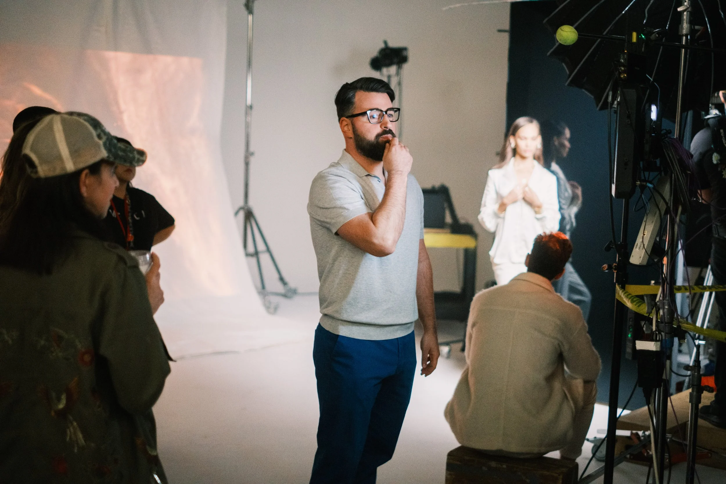 A man with glasses, a beard, and a gray polo shirt appears to be thinking or observing on a photoshoot set. There are people and equipment around him, with a woman in white in the background and others on the set.