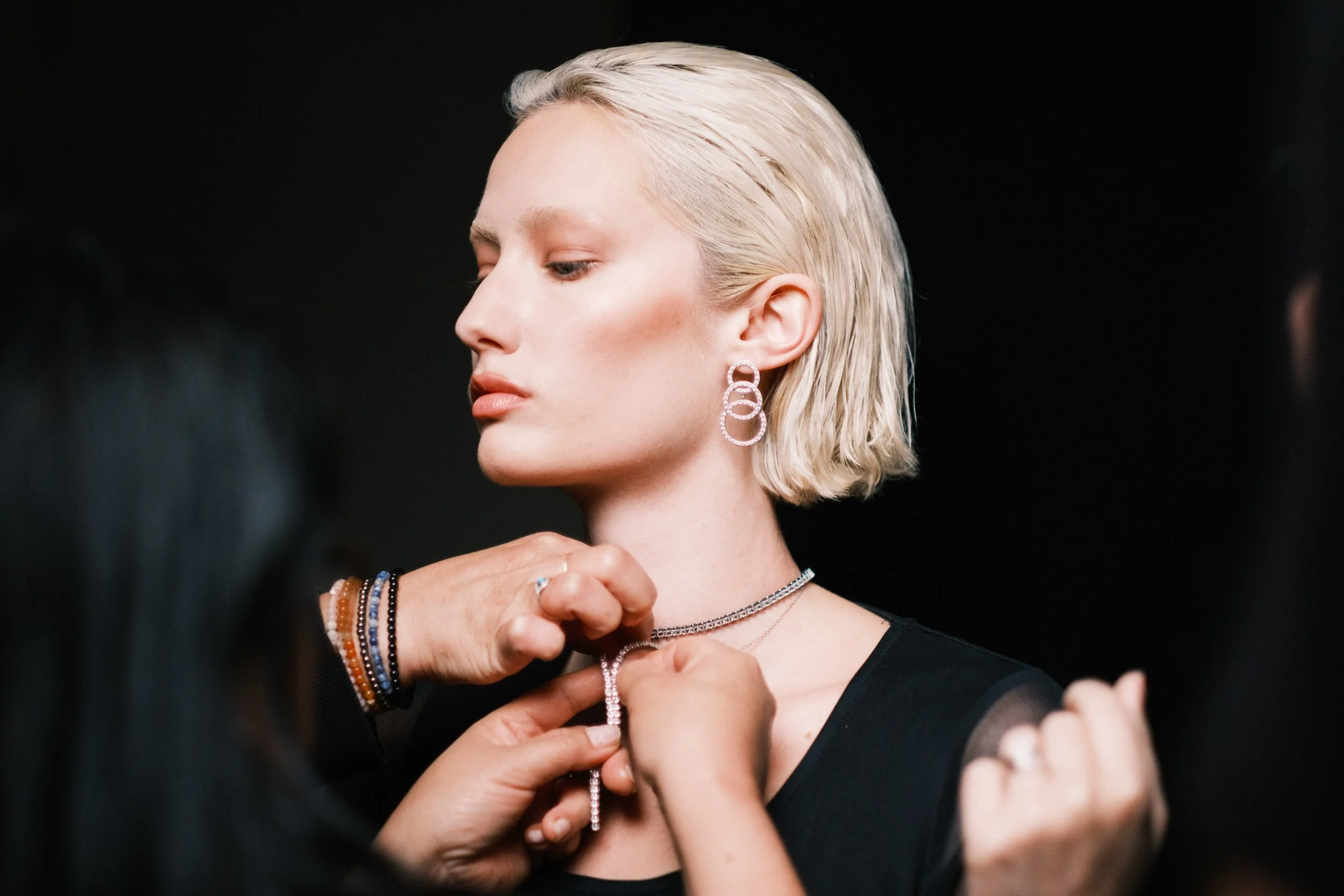 A woman with platinum blonde hair and light skin wearing silver jewelry is being assisted with a necklace. She is dressed in black and stands against a dark background. Branded entertainment director and showrunner creating scripted/unscripted films