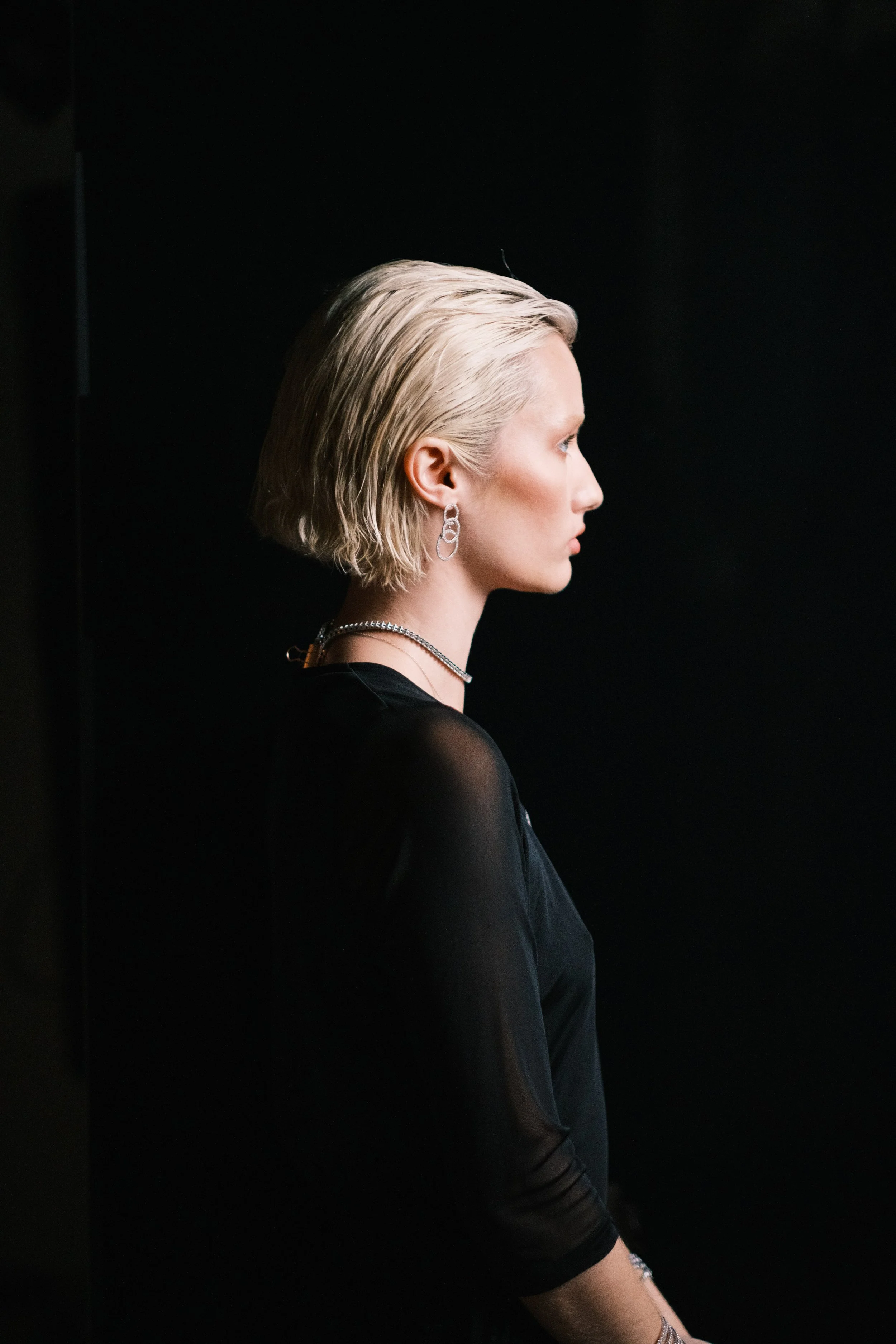 Side profile of a woman with short platinum blonde hair, wearing earrings, a necklace, and a black top with sheer sleeves, standing against a black background.
