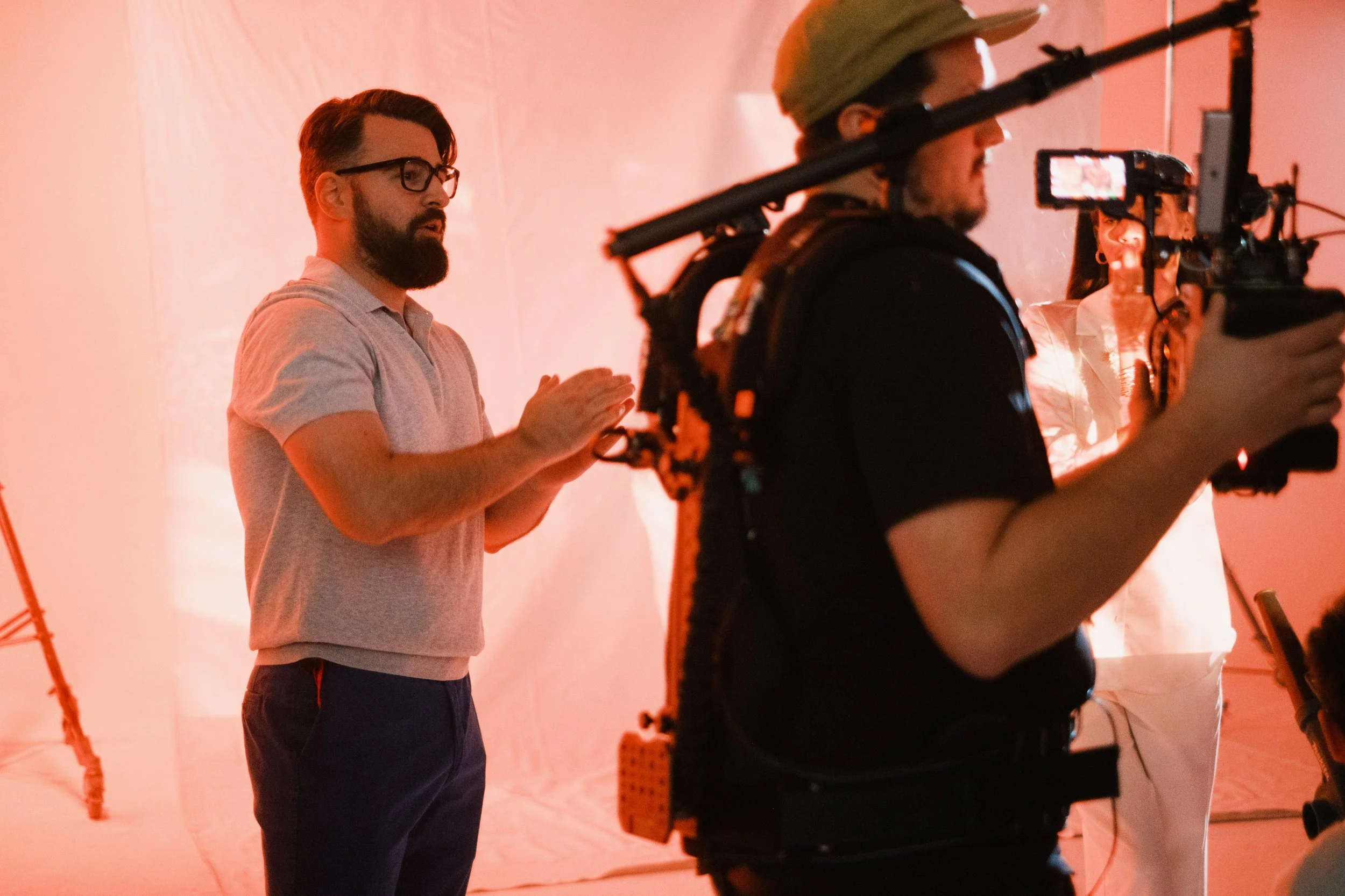 People working on a film or video production set with camera operators, one man with glasses and a beard, another person in a white outfit, against a pinkish background.