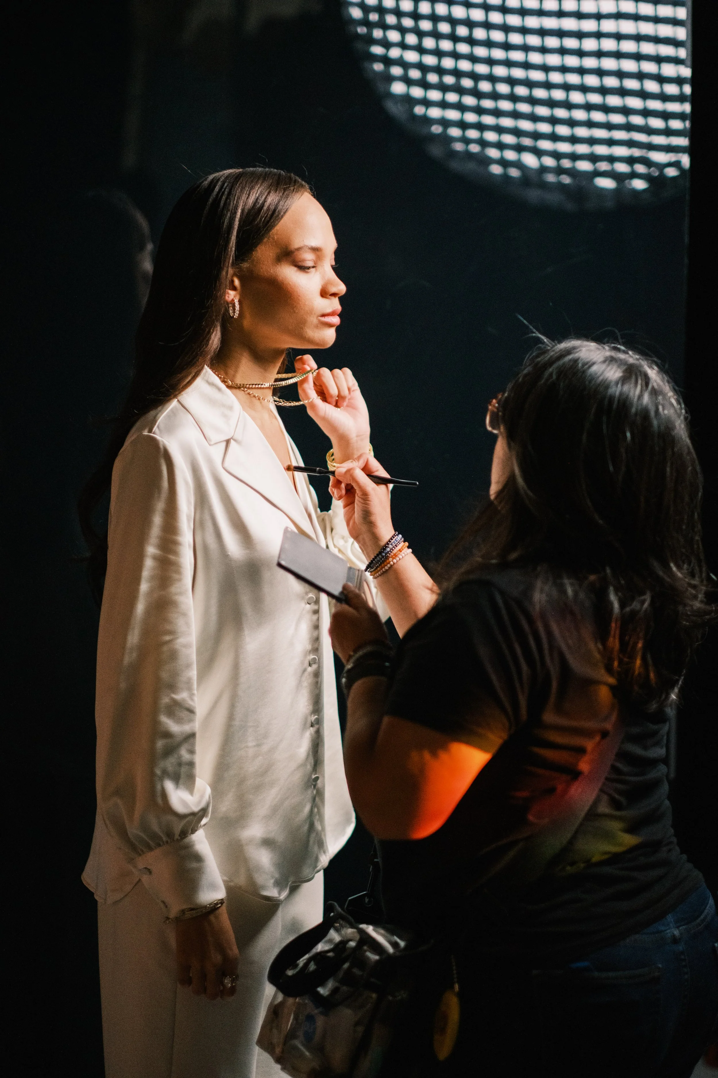 A woman in a satin white blouse and matching pants is getting her makeup or face styled by a makeup artist or stylist. The stylist is holding a makeup brush and a small palette or mirror, with a professional lighting setup visible in the background.