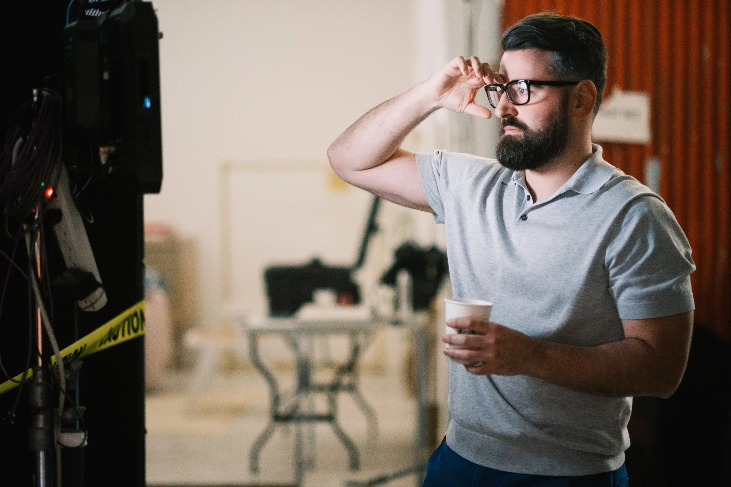 A man with glasses and a beard, wearing a gray polo shirt, adjusting his glasses with one hand and holding a paper cup in the other, standing indoors near a server or electronic equipment.