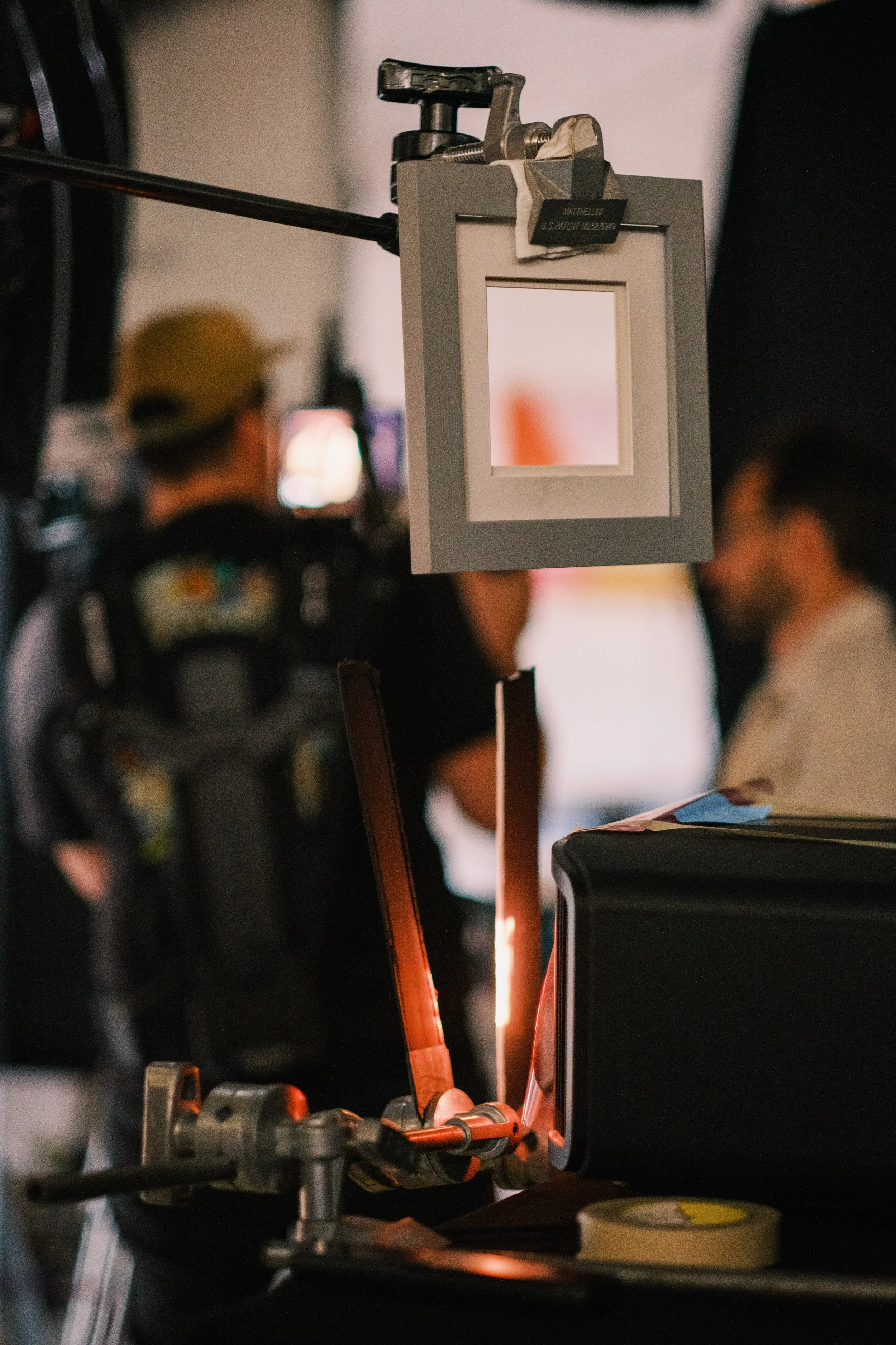 A metal frame with a small display window is mounted on a clamp, with a laser or lighting device shining on a surface below, in a studio or production setting with people and equipment in the background.