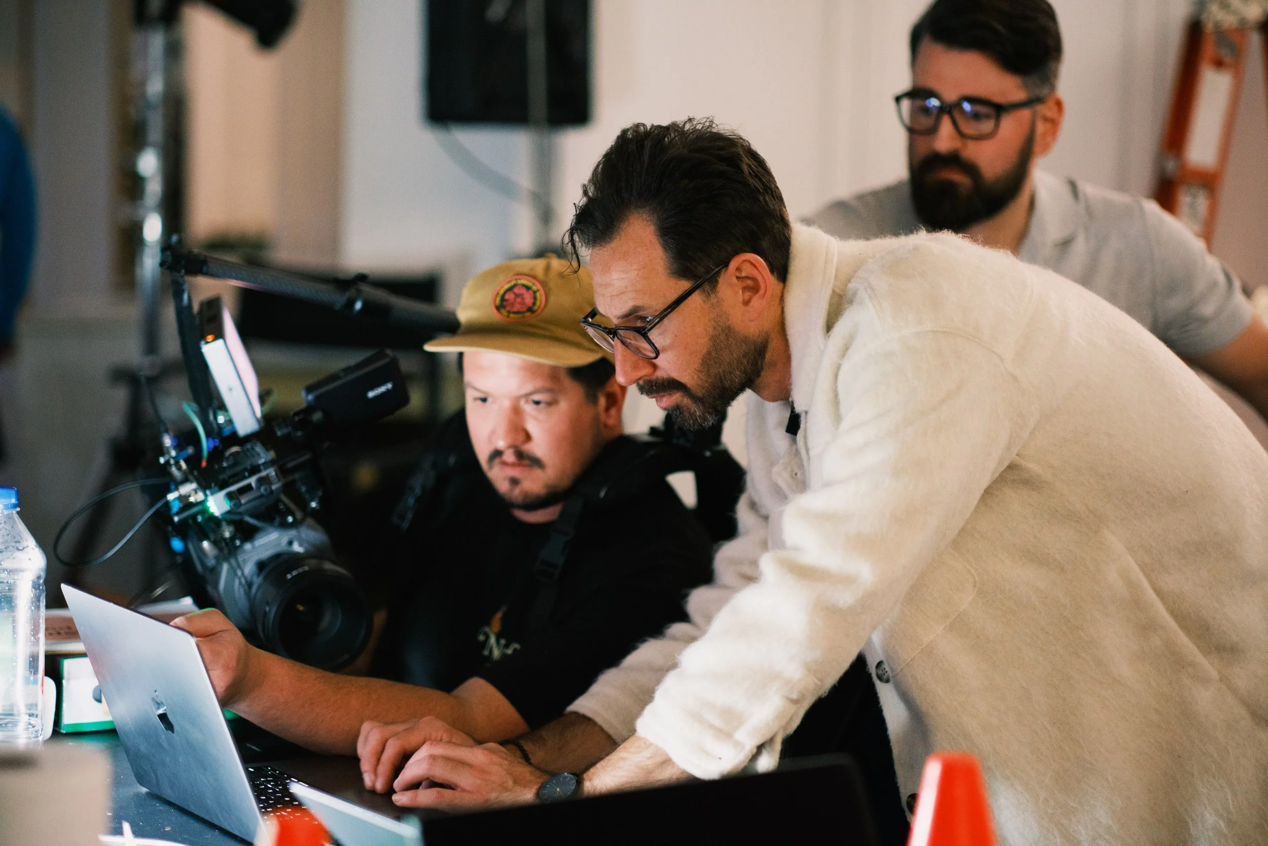 Three men working together on a film set, looking at a laptop screen. One man is operating a camera, and another is wearing a beige hat and glasses. They are in a busy indoor environment.