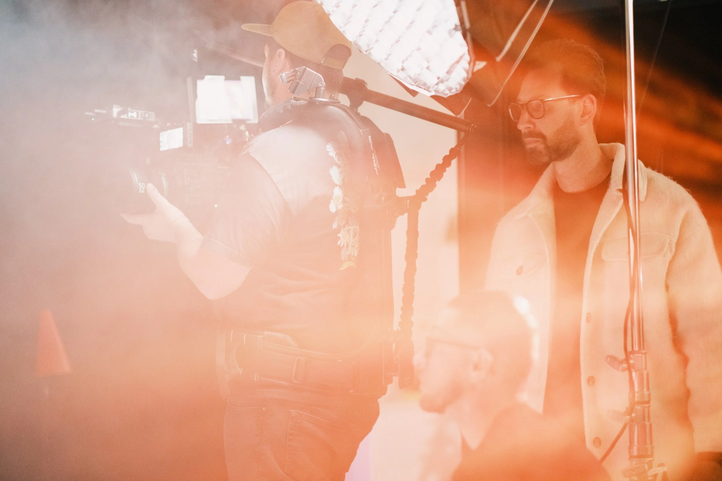 A behind-the-scenes photo showing a film crew preparing to shoot a scene, with a man wearing glasses standing next to a camera and a crew member operating equipment, illuminated by bright lights and orange-tinted lighting.