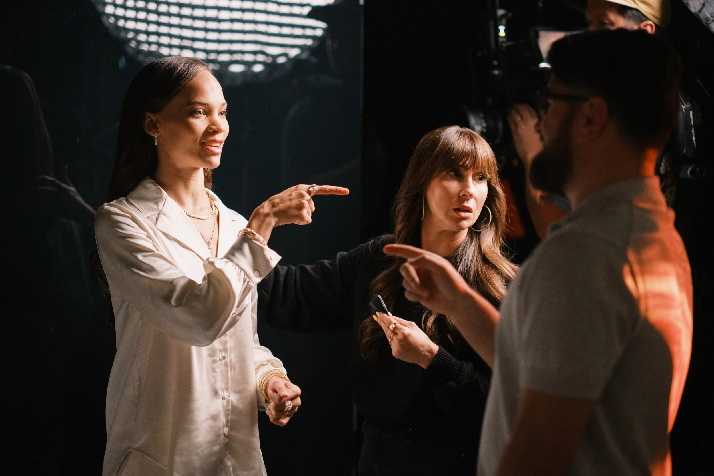 Three people engaged in a discussion, with two women and one man, in a dimly lit setting. One woman is pointing and speaking, while the other woman and the man listen, with a media camera in the background.
