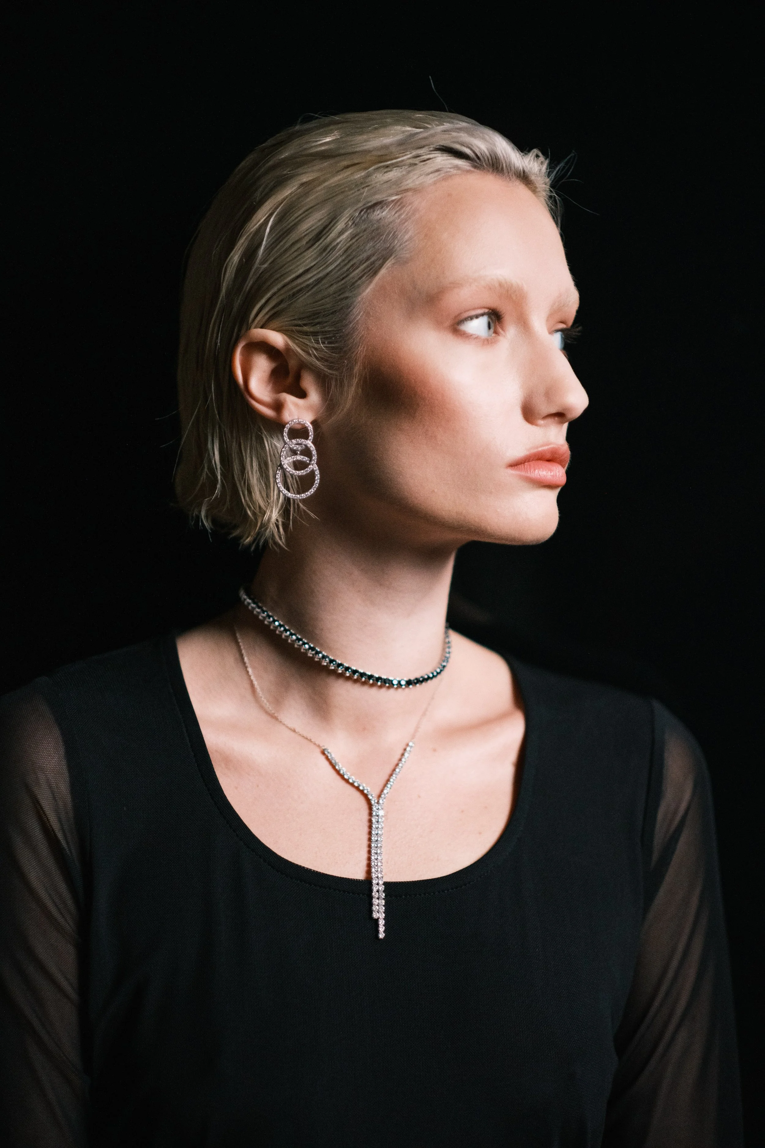 A woman with short blonde hair wearing layered earrings, a choker, and a lariat-style necklace, dressed in a black top with sheer sleeves, posing against a dark background.