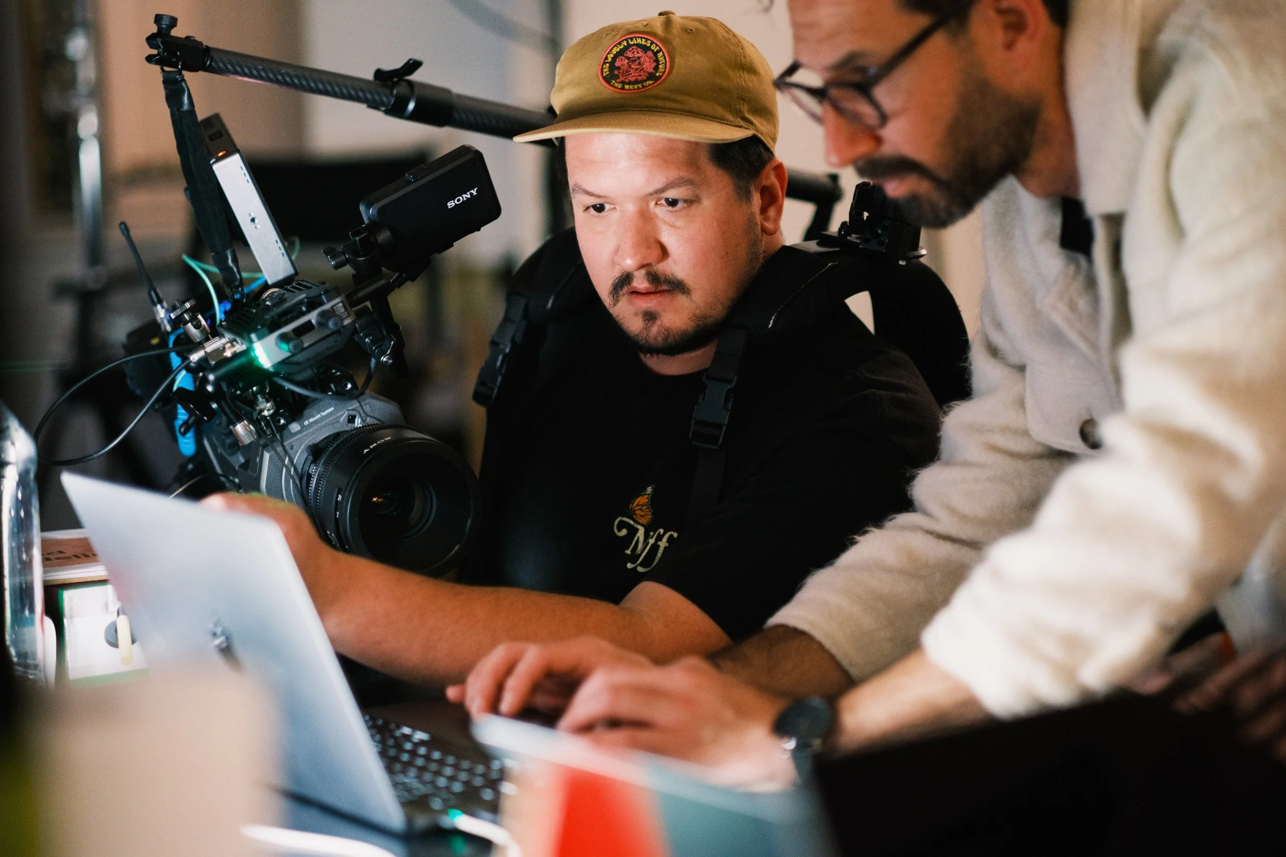 Two men working on a film set; one man holding a camera, the other man typing on a laptop.