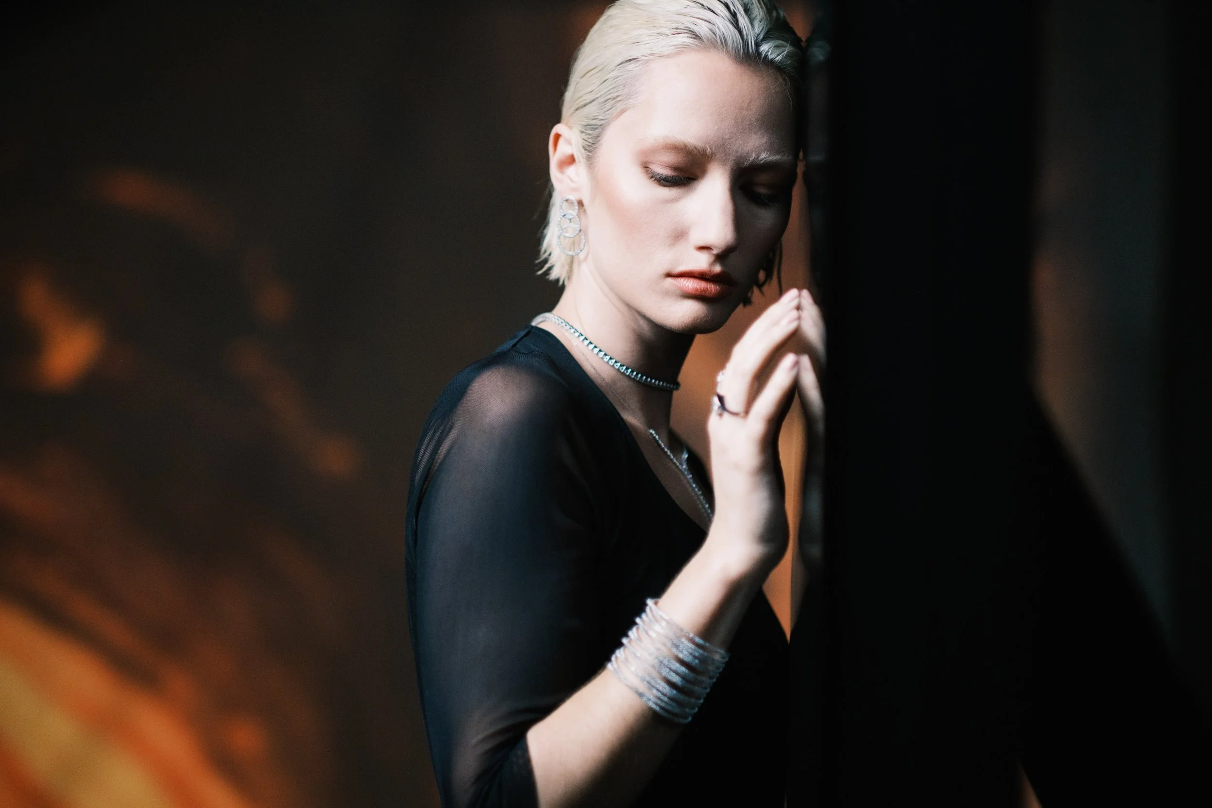 A woman with short platinum blonde hair, wearing a black sheer top, is touching a dark wall with her eyes closed, jewelry including earrings, necklaces, rings, and bracelets, in a dimly lit setting.
