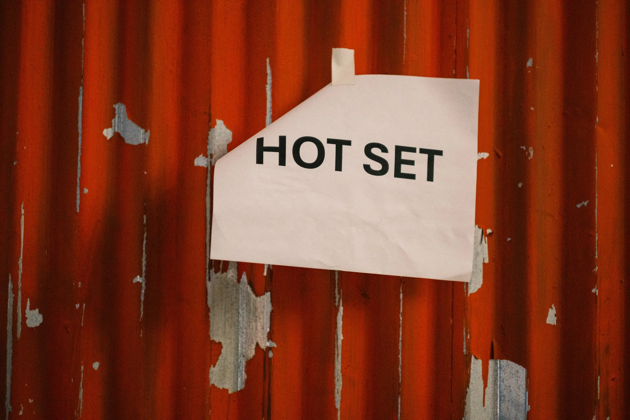 Branded entertainment director and showrunner creating scripted/unscripted films and campaigns for brands. U.S.-based, global work.White paper sign with bold black letters reading "HOT SET" taped to a rusty, corrugated metal wall.