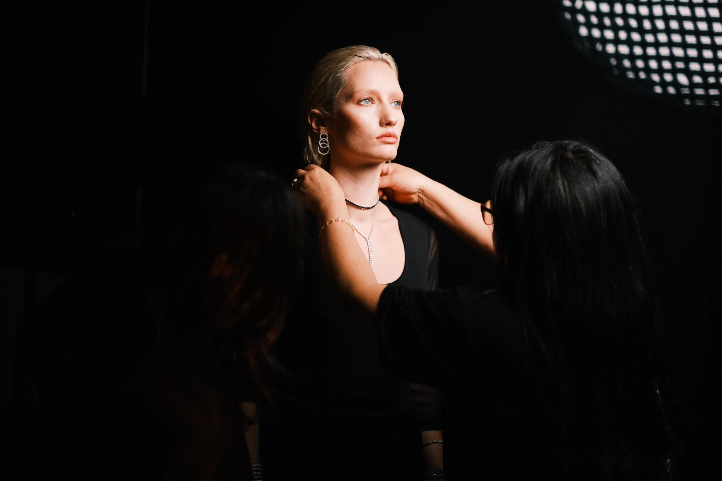 A blonde woman with jewelry sits posed while two women assist her in preparing for a photoshoot or event, set against a dark background with stage lighting.