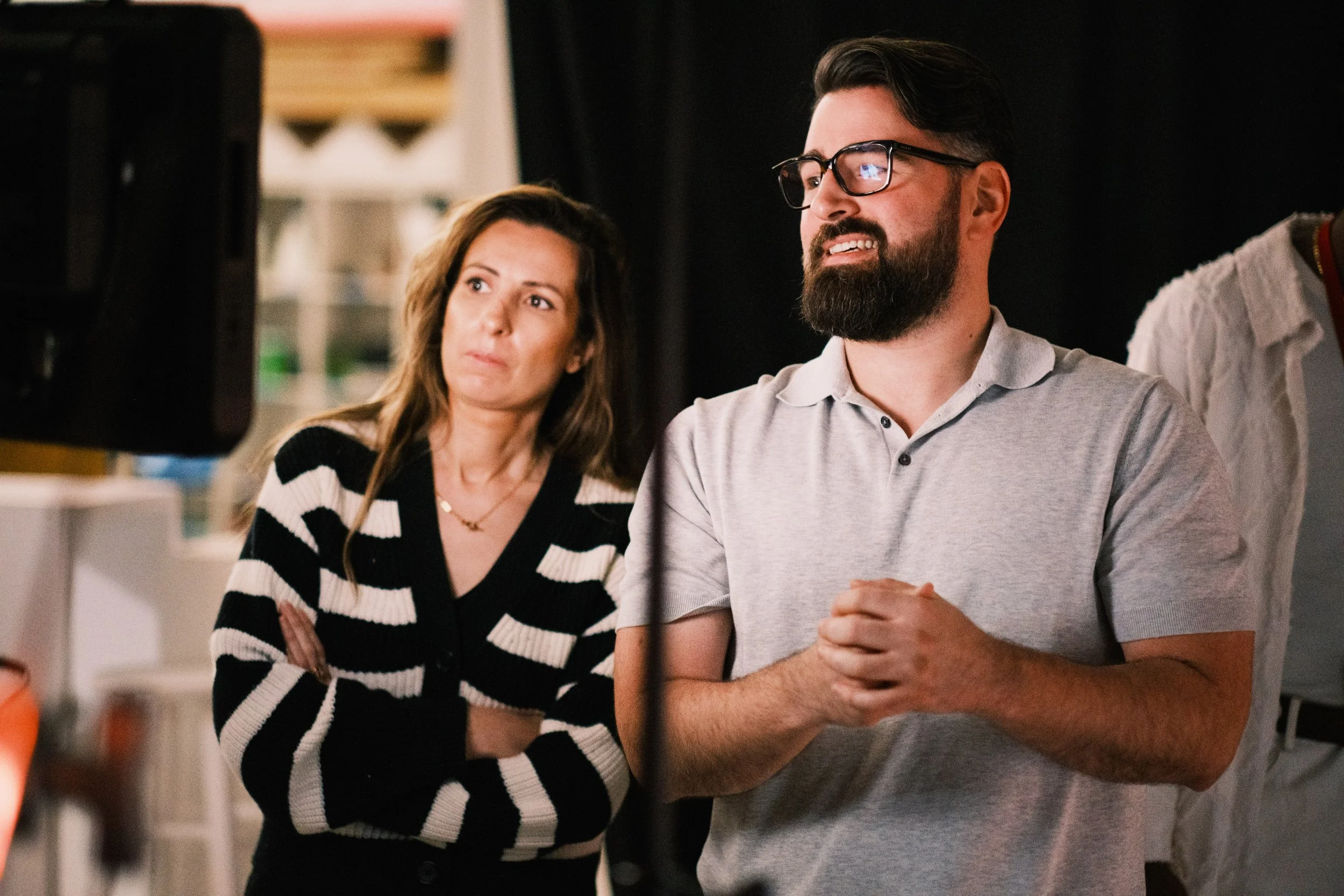 Two adults watching a screen or presentation, with the man wearing glasses and a light polo shirt, clasping his hands, and the woman in a black and white striped blazer with arms crossed, seeming attentive and thoughtful.