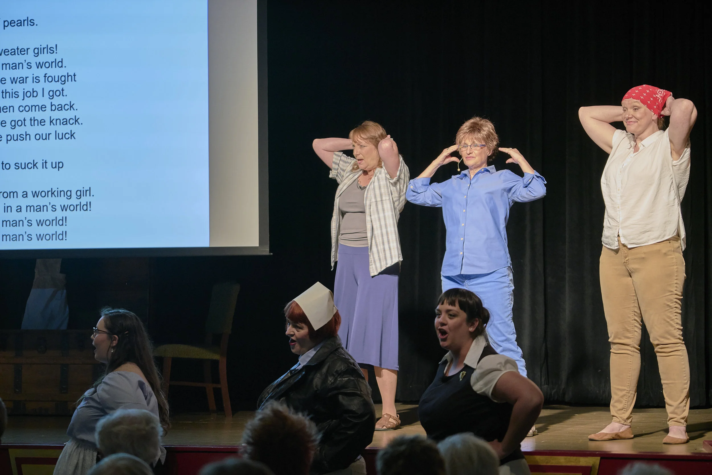 L to R on stage, Lisa Green as Dorothea Lange, Darla Hoffmann as Narrator, and Liz Vincent as Adeline Van Buren; and L to R below stage, Brooke Sauter as Hazel Funk Holmes, Stephanie Rhein as Edna, a Phillips 66 Highway Hostess, and Karen Wright as Ray Eames sing “Bandanas tied around our curls” in the original song, “Making Good in a Man’s World,” the finale to Act One in the Pontiac production June 22-23. (Photo by Mitchell T. Fransen, courtesy of the City of Pontiac.)