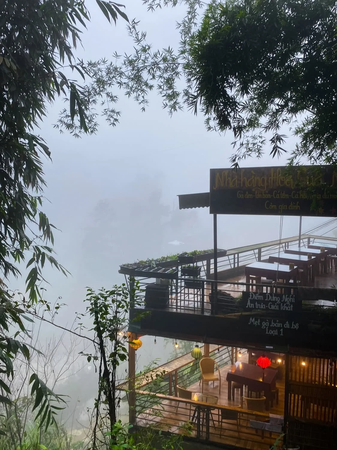 A cozy hillside restaurant in Cat Cat Village sits on stilts surrounded by mist and bamboo, with signs in Vietnamese and hanging lanterns creating a warm atmosphere.