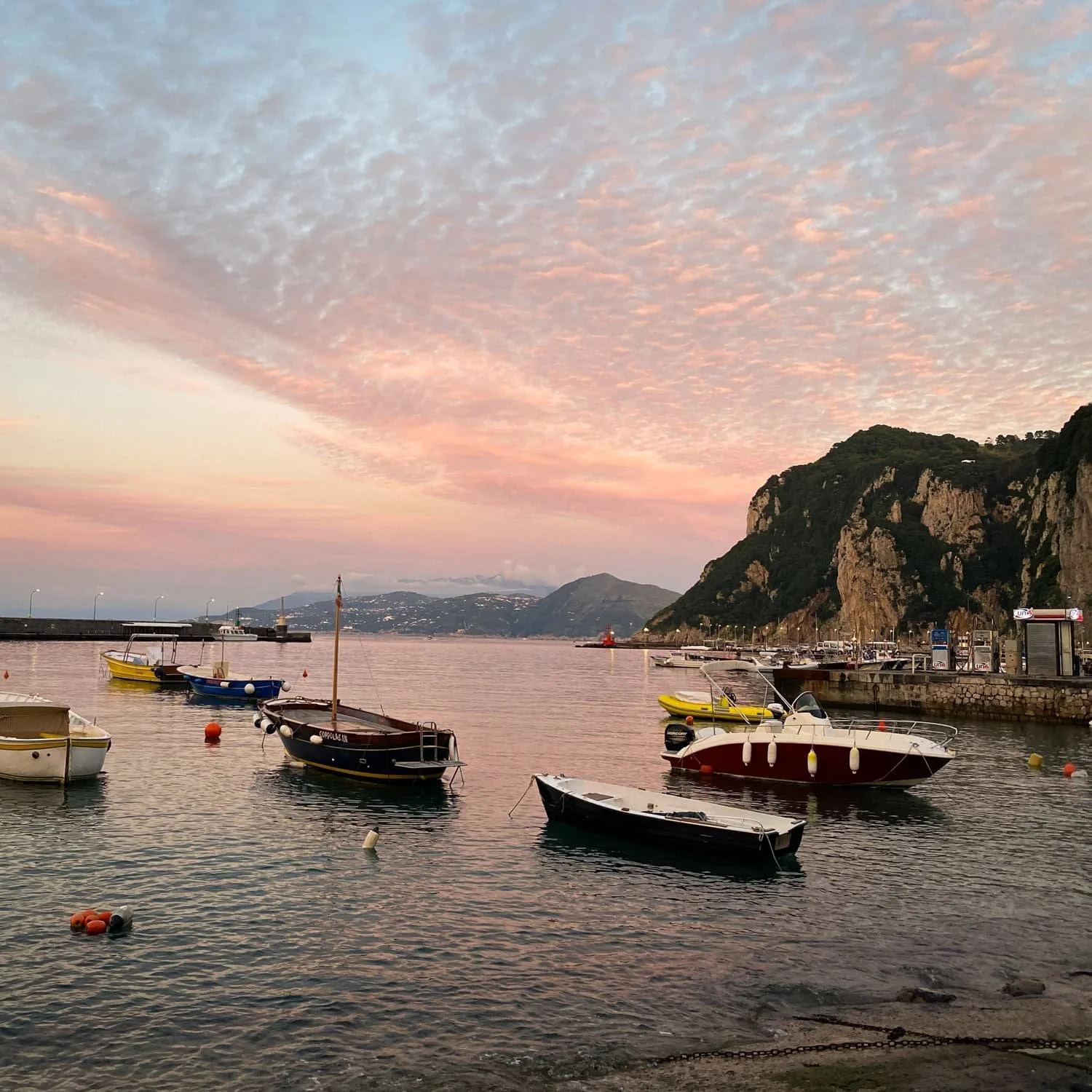 Small boats floating in a calm harbor at sunset with pink clouds reflecting on the water and cliffs rising along the shoreline. Watching sunset by the harbor is one of the relaxing Amalfi Coast things to do.