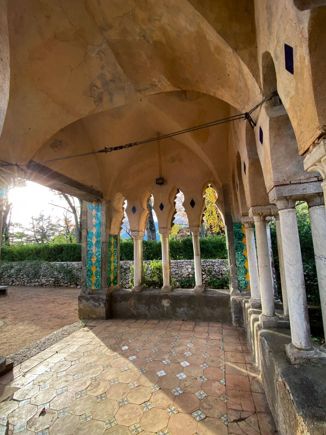 Sunlight streams through an open stone pavilion with arched columns, patterned floor tiles, and colorful mosaic accents in Ravello. The historic architecture showcases one of the cultural things to do in Ravello.