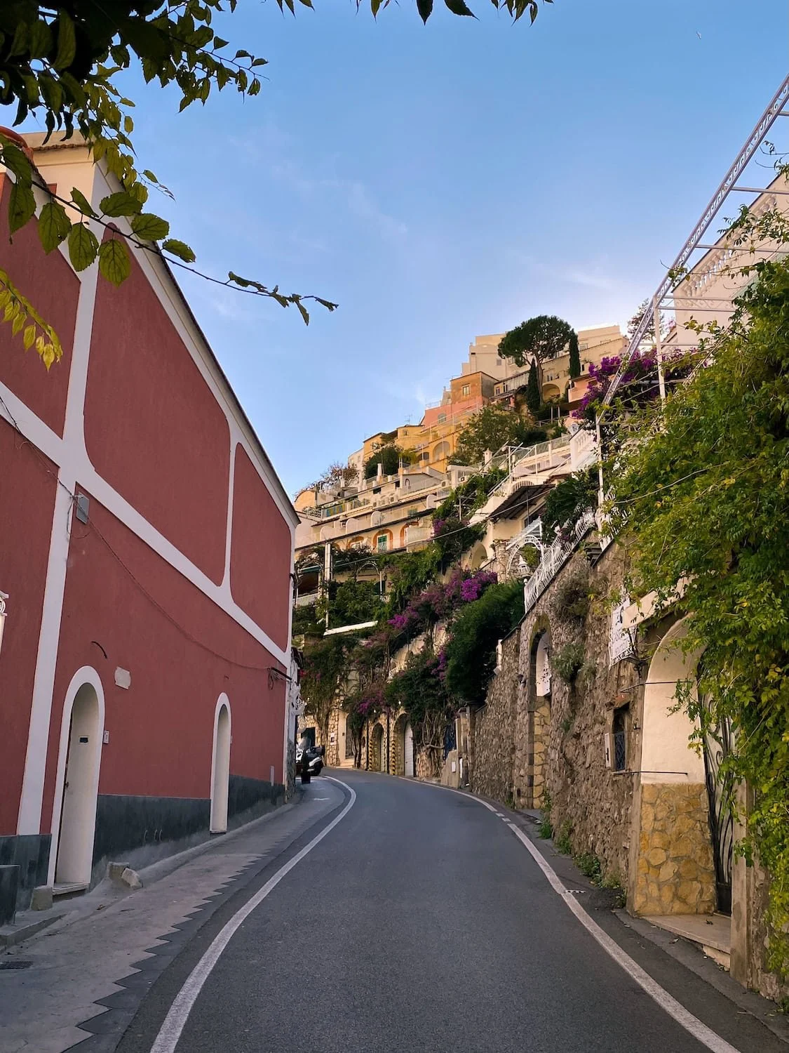 Curving coastal road in Positano lined with colorful cliffside homes, stone walls, and flowering plants. The quiet street winds through the hillside town at sunset.