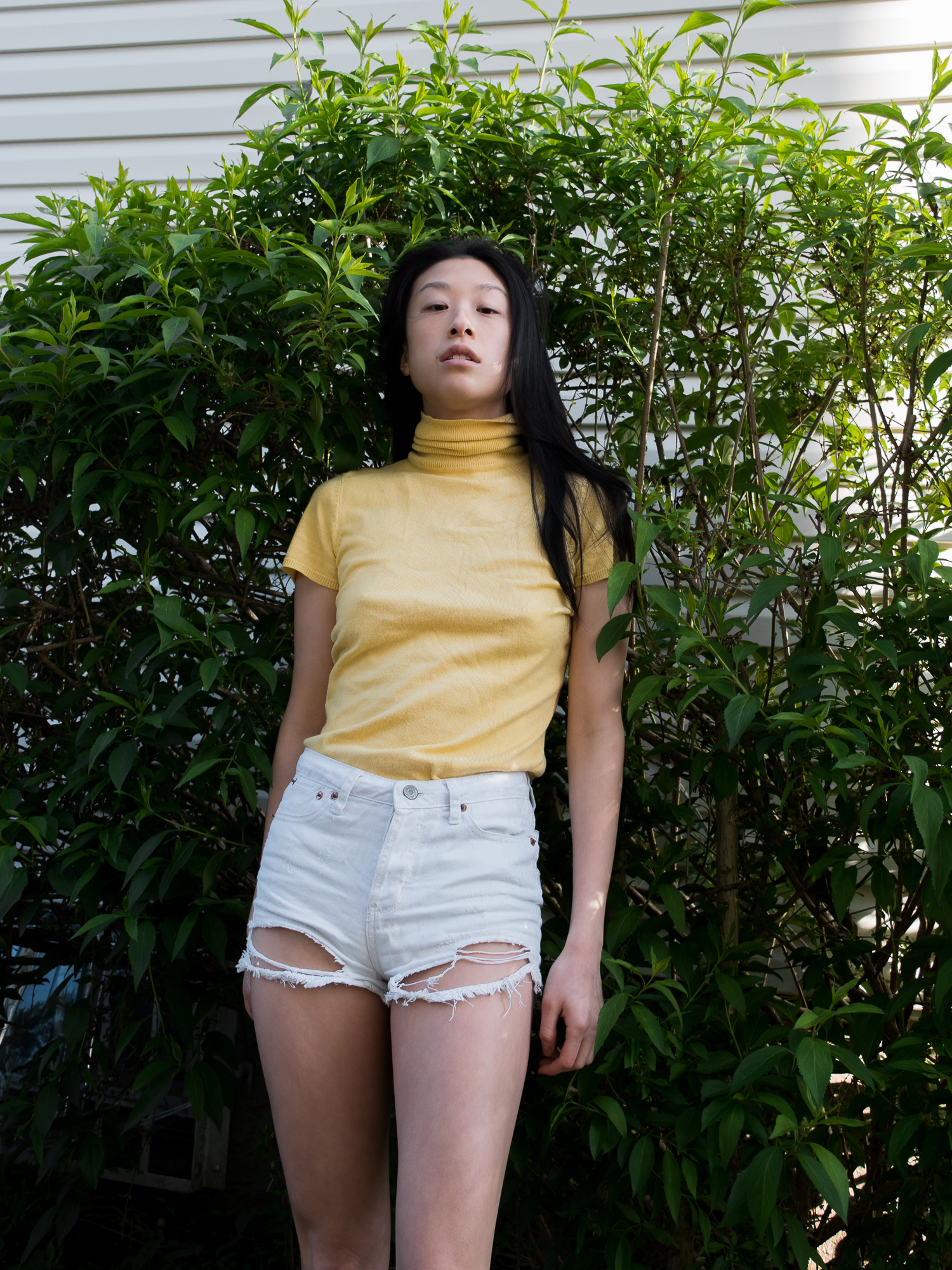 Close-up of female blogger in Yellow short-sleeved turtleneck and distressed white shorts against green leafy background