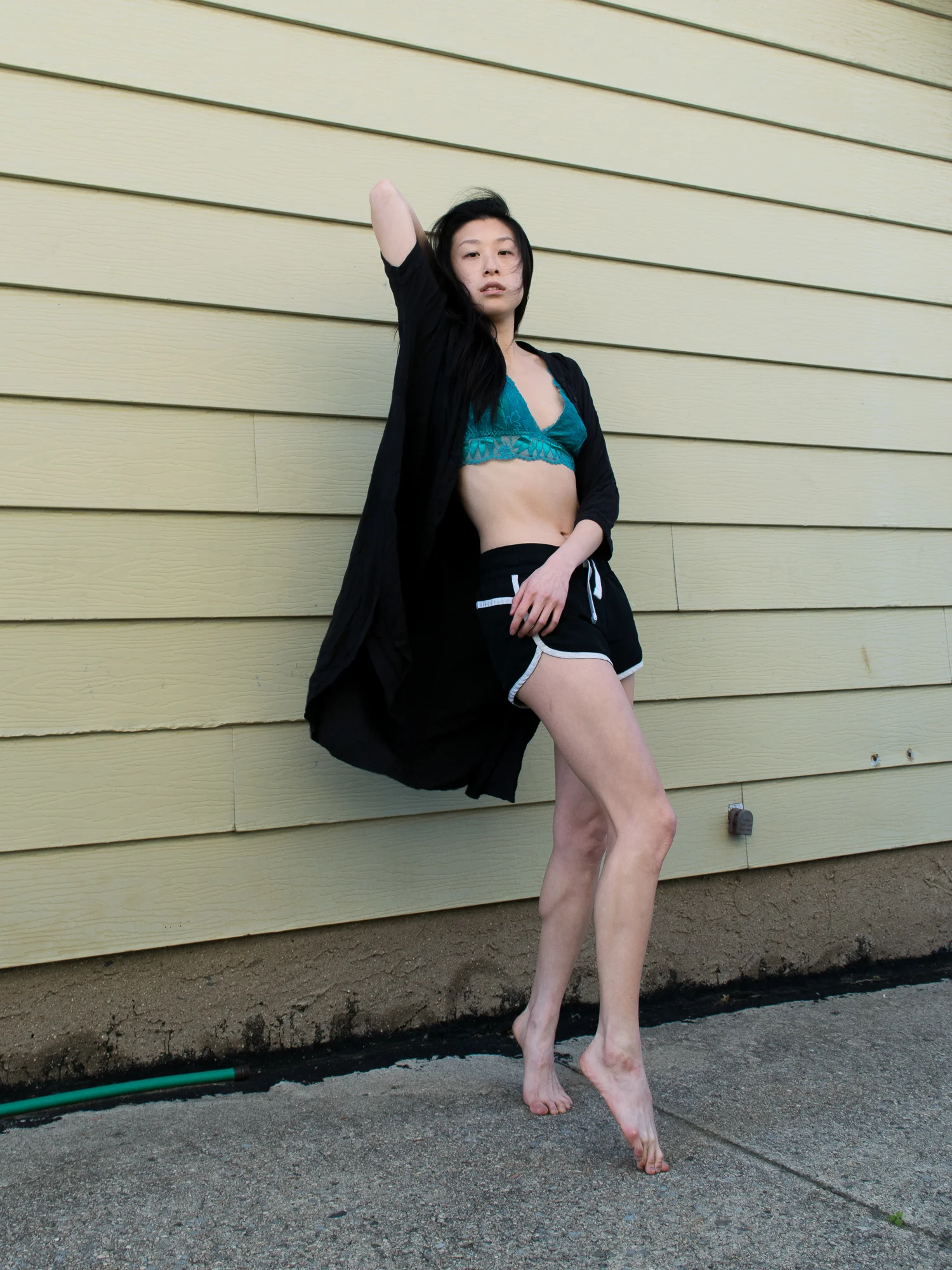 Female east asian style blogger wearing bright blue bralette top, black and white athletic shorts, black shirt-dress leaning against a yellow wall