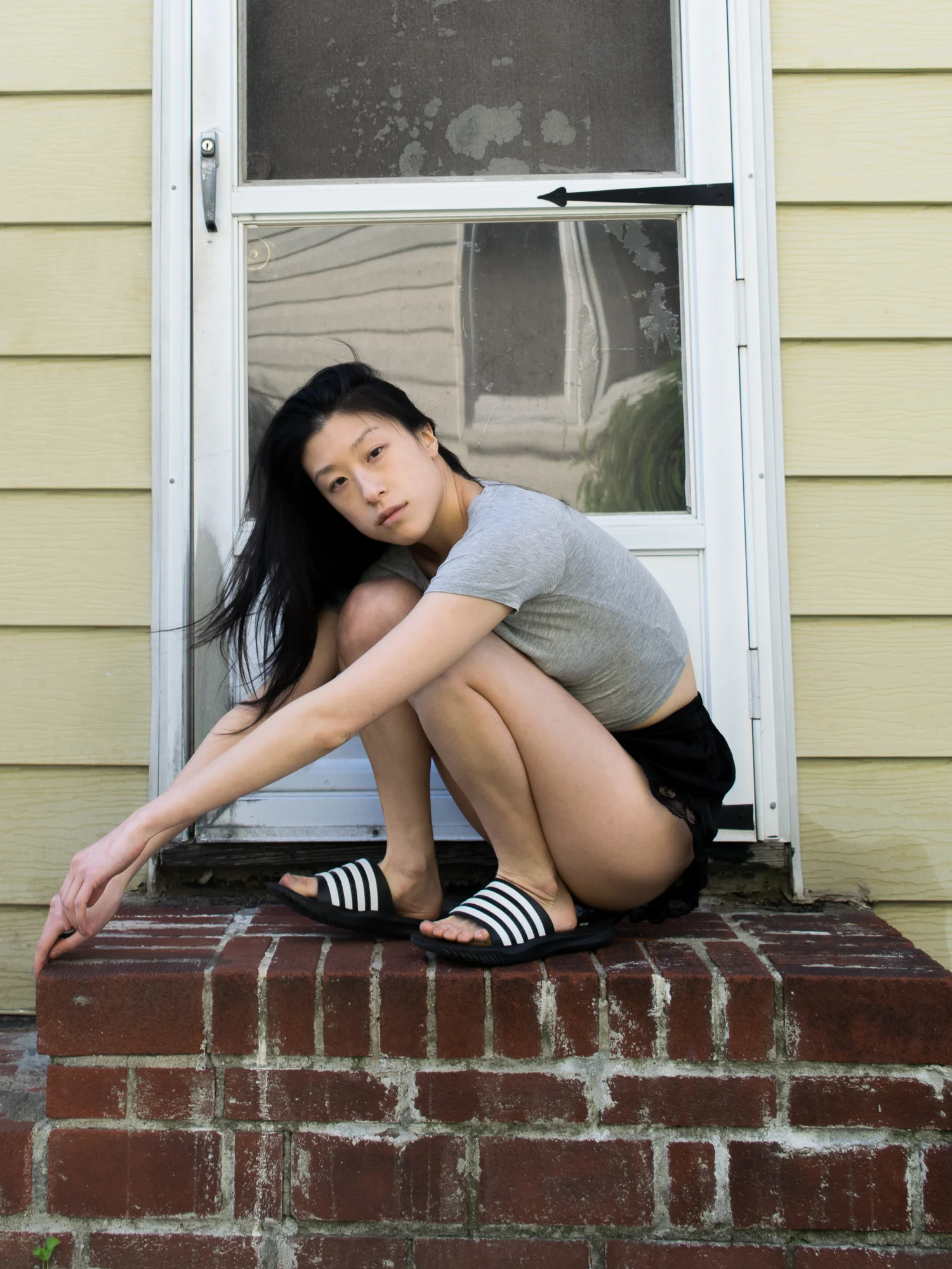 Female, east asian blogger in grey cropped t-shirt, lacy black shorts, black and white striped sandals, posing on brick stoop
