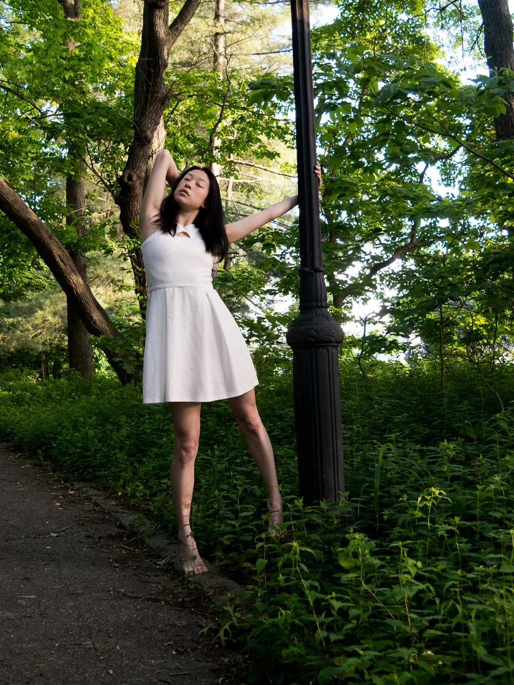 Female east asian style blogger wearing white a-line dress holding on to lamp post in park