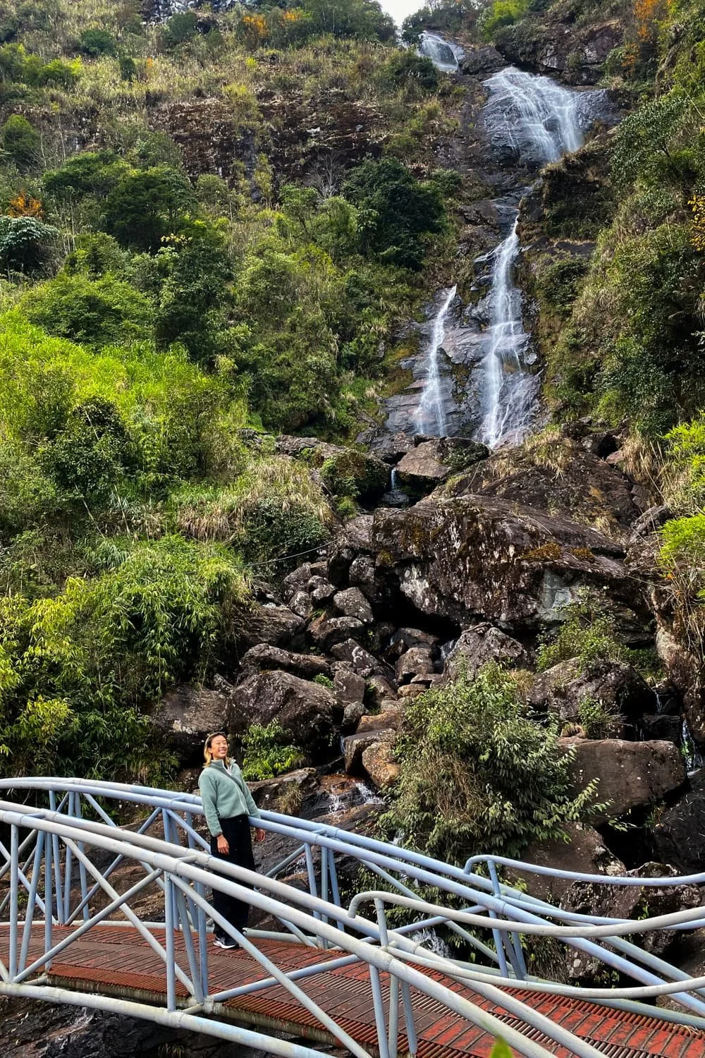  A person stands on a curved metal bridge in front of a multi-tiered waterfall cascading down a rocky mountain slope. Visiting scenic waterfalls like this is a refreshing adventure on a Sapa itinerary. 