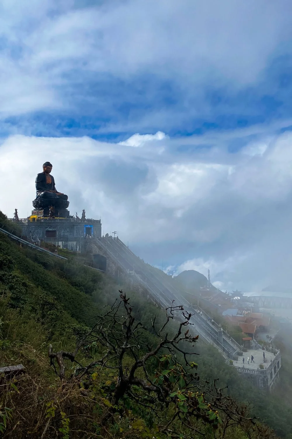 A large bronze Buddha statue sits serenely on a mountaintop platform surrounded by cloud-covered peaks and stone staircases. Spiritual landmarks like this make Fansipan a must-see in a Sapa itinerary. 