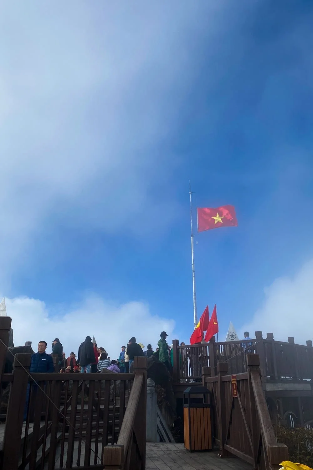  Tourists gather on the wooden viewing platform at the summit of Fansipan, the highest peak in Vietnam, under the waving red national flag. Reaching this iconic mountaintop is a bucket list moment on any Sapa itinerary. 