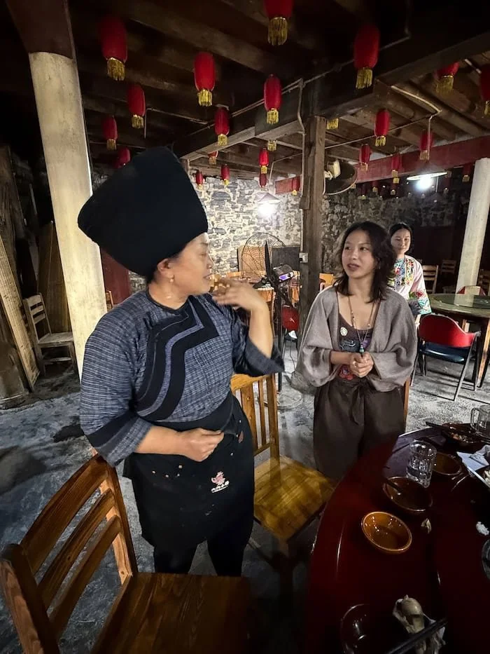 A Miao restaurant owner sharing stories with travelers inside her restaurant in a traditional village.