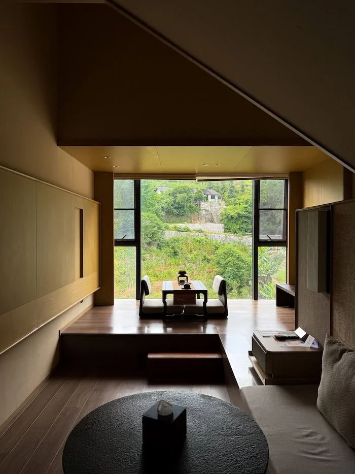 Modern hotel room with a large window overlooking lush green hills and village scenery.