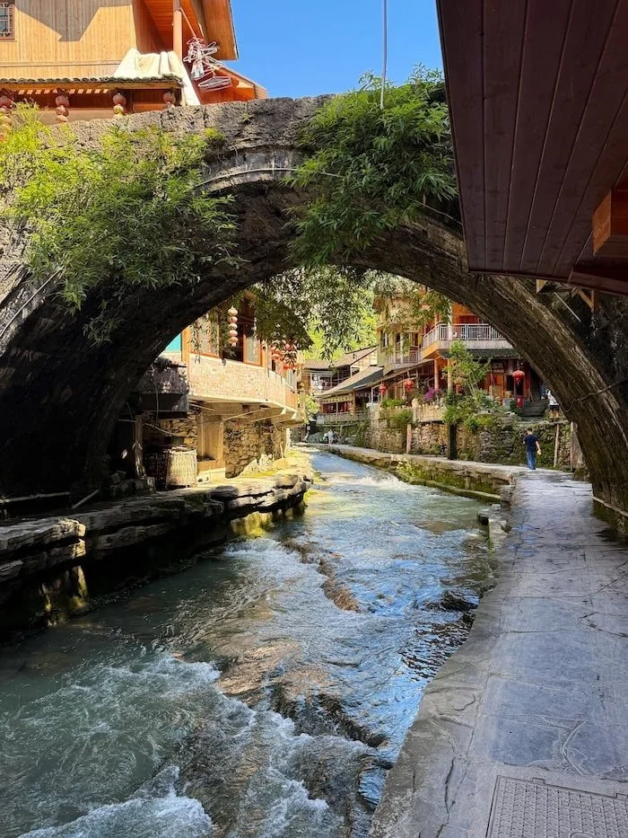 Stone arch bridge spanning a river through a historic village in China.