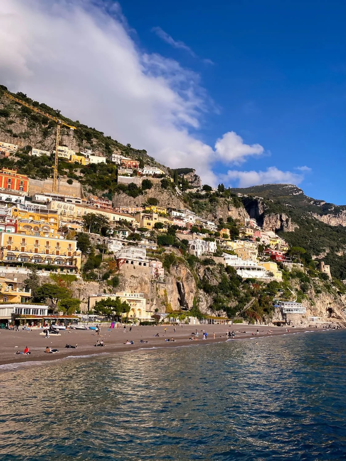 Wide view of Positano beach with colorful cliffside homes rising above the shoreline along the Amalfi Coast. Calm blue water and dramatic clouds surround the seaside town on a sunny afternoon.